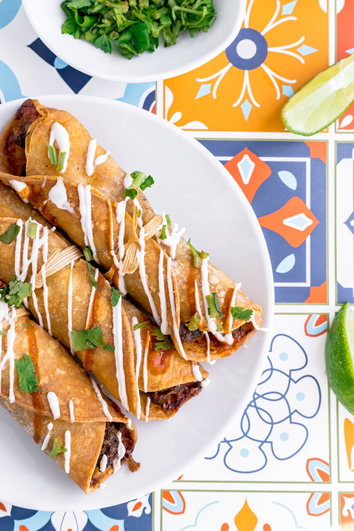 Three crispy rolled tacos, just like air fryer beef taquitos, topped with white sauce and cilantro, served with limes on a colorful tile table—a perfect gluten free Mexican dinner.