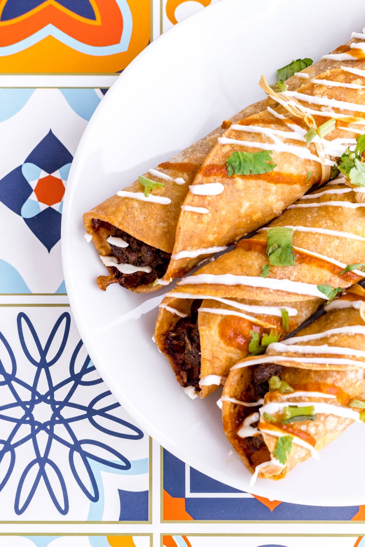 Three crispy rolled tacos topped with sour cream and cilantro on a white plate, with a colorful tile background—this easy taquito recipe brings the perfect gluten free Mexican dinner to your table.