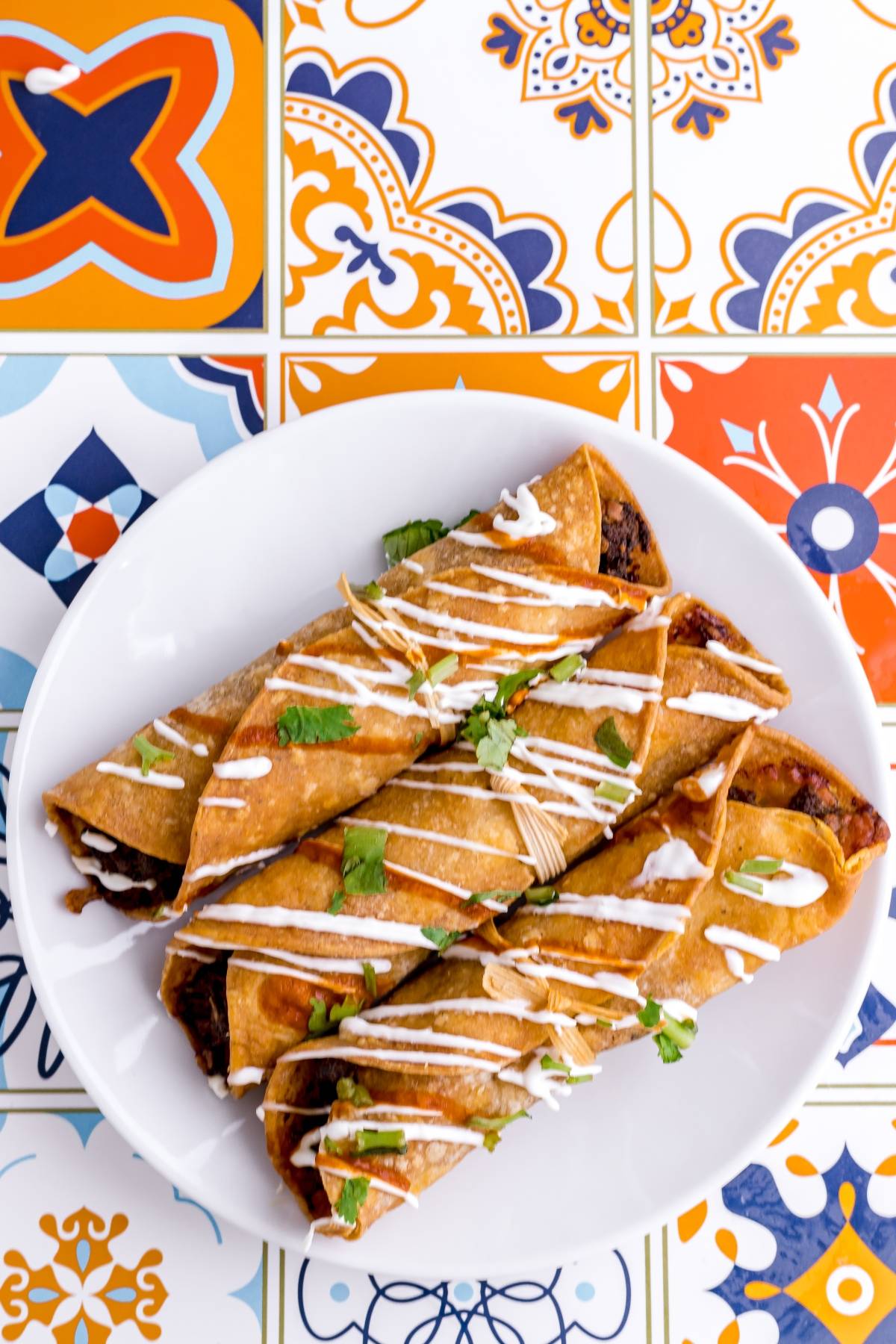 Four crispy air fryer taquitos topped with crema and cilantro on a white plate, set on colorful patterned tiles—a delicious and easy taquito recipe for your gluten free Mexican dinner.