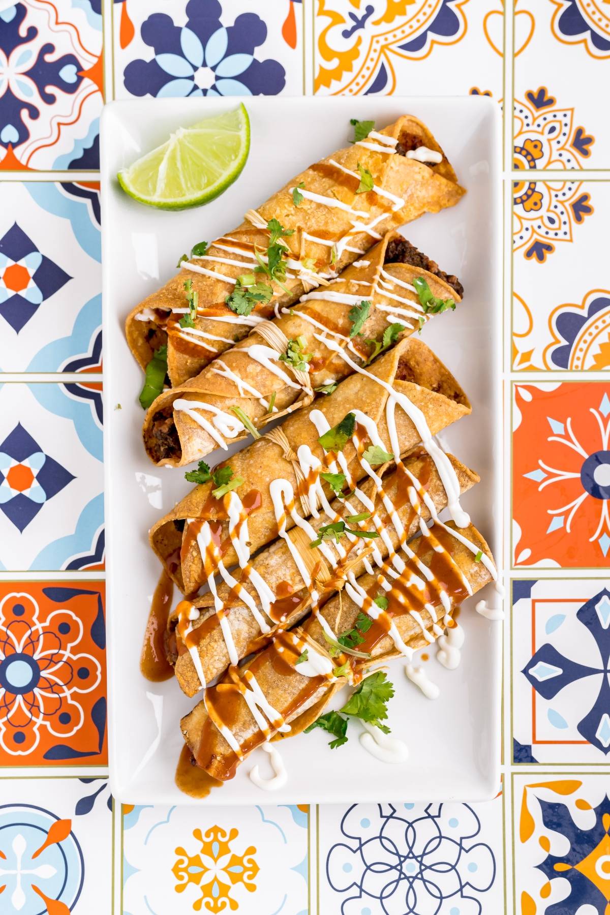 Four crispy air fryer taquitos on a plate, topped with sauce, cream, cilantro, and a lime wedge, rest on colorful tiles—a true contender for the best air fryer recipe.