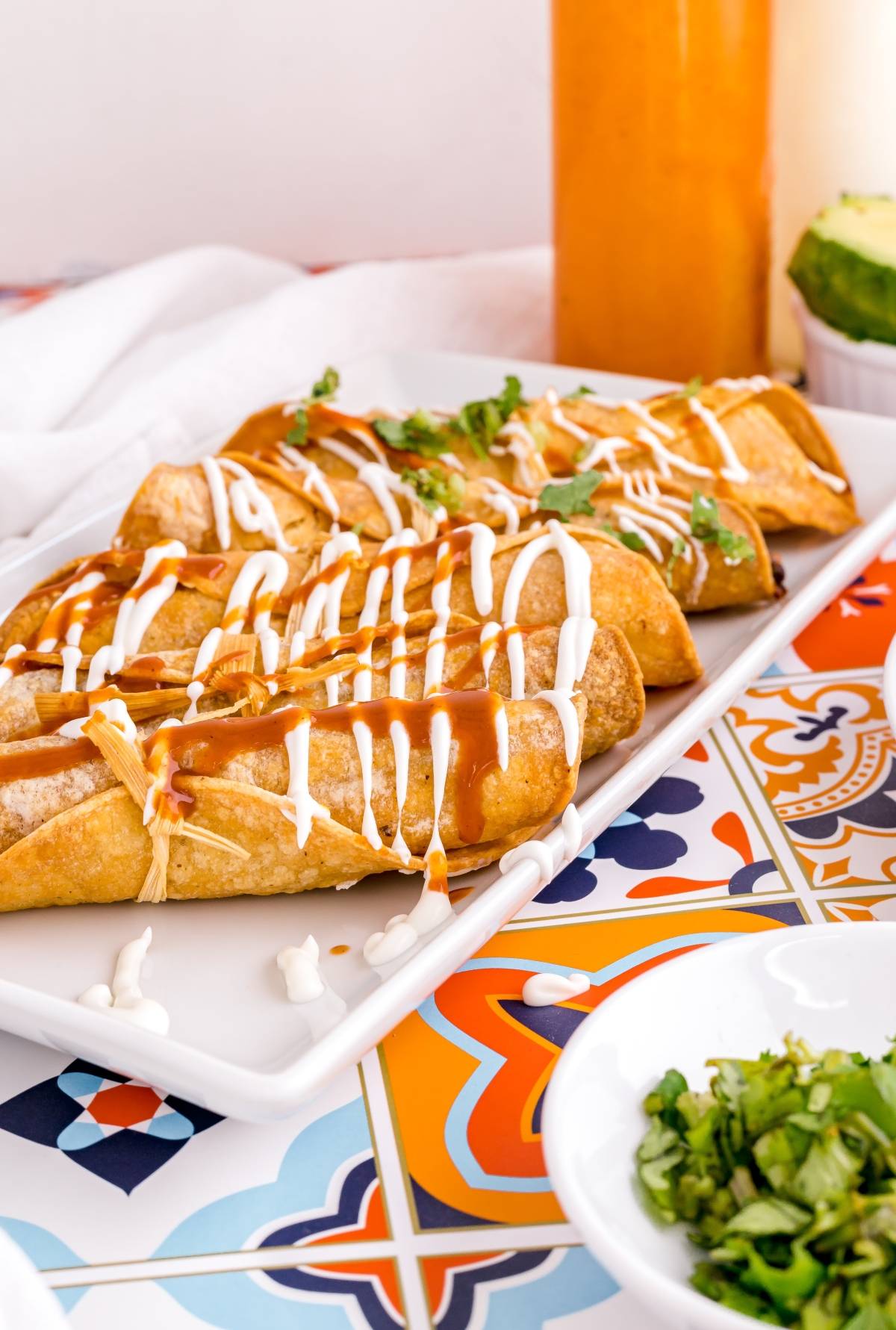 Five crispy air fryer beef taquitos drizzled with sour cream and sauce, garnished with cilantro, served on a colorful tiled table—perfect for a gluten free Mexican dinner.