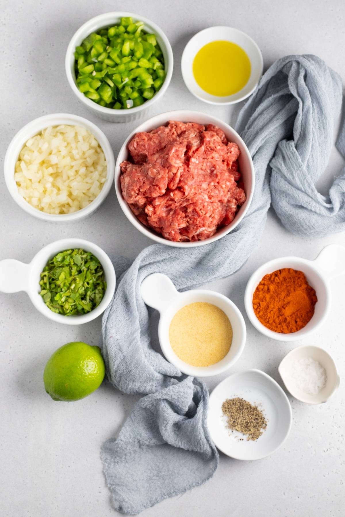 Bowls of ground meat, chopped onion, green pepper, spices, oil, lime, and herbs on a gray surface—perfect for preparing gluten free Mexican dinner favorites like air fryer taquitos.