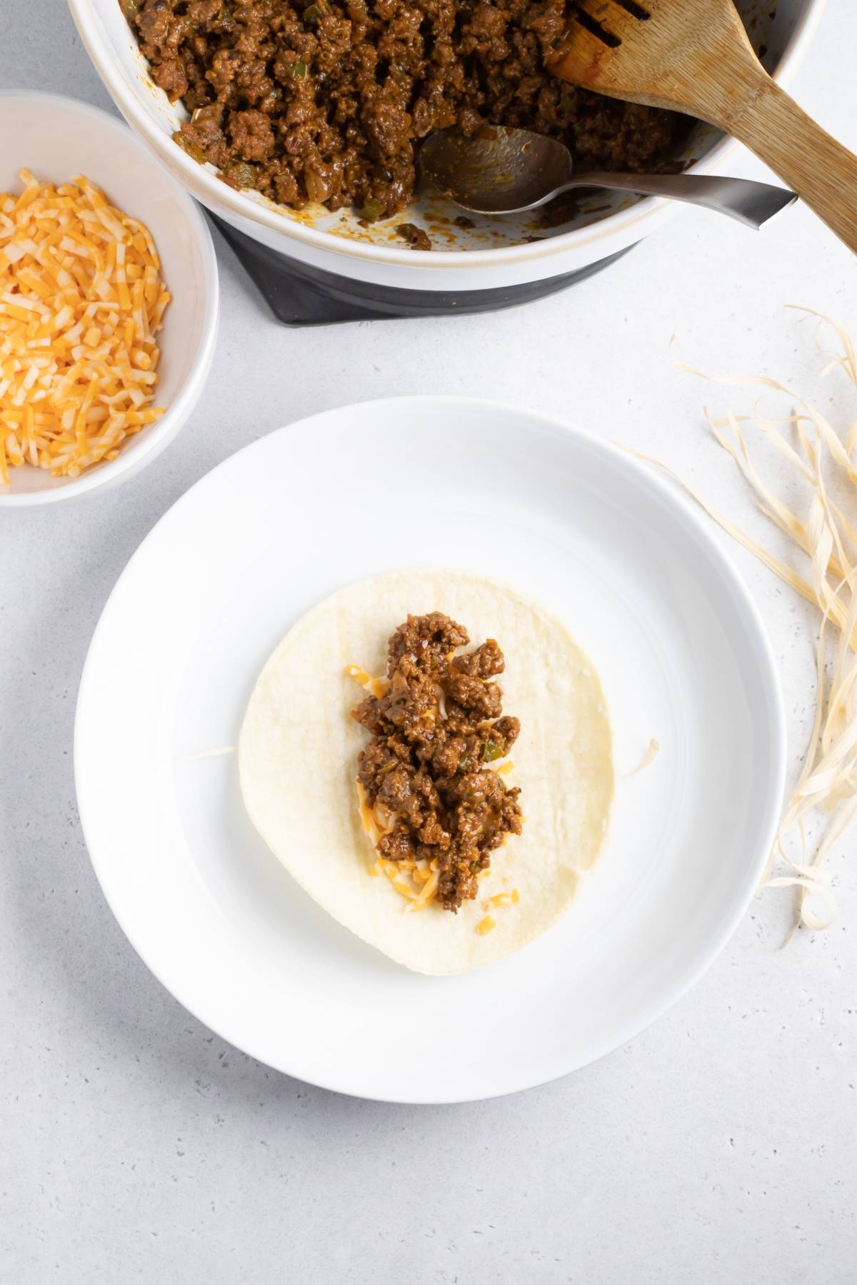 A tortilla on a plate is topped with ground beef filling, making it perfect for an easy taquito recipe. Cheese and more beef in bowls nearby make this the best air fryer recipe for a quick and tasty meal.