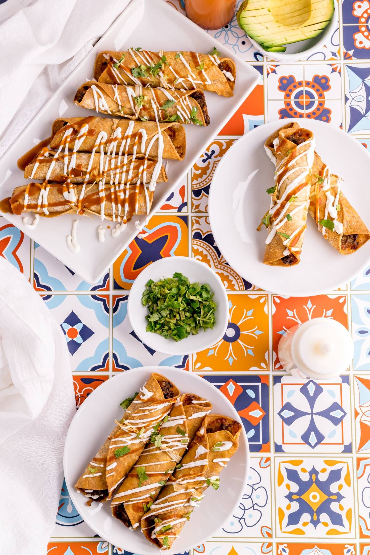 Plates of crispy air fryer beef taquitos drizzled with crema, served with chopped greens on a colorful tiled table—your new best air fryer recipe.