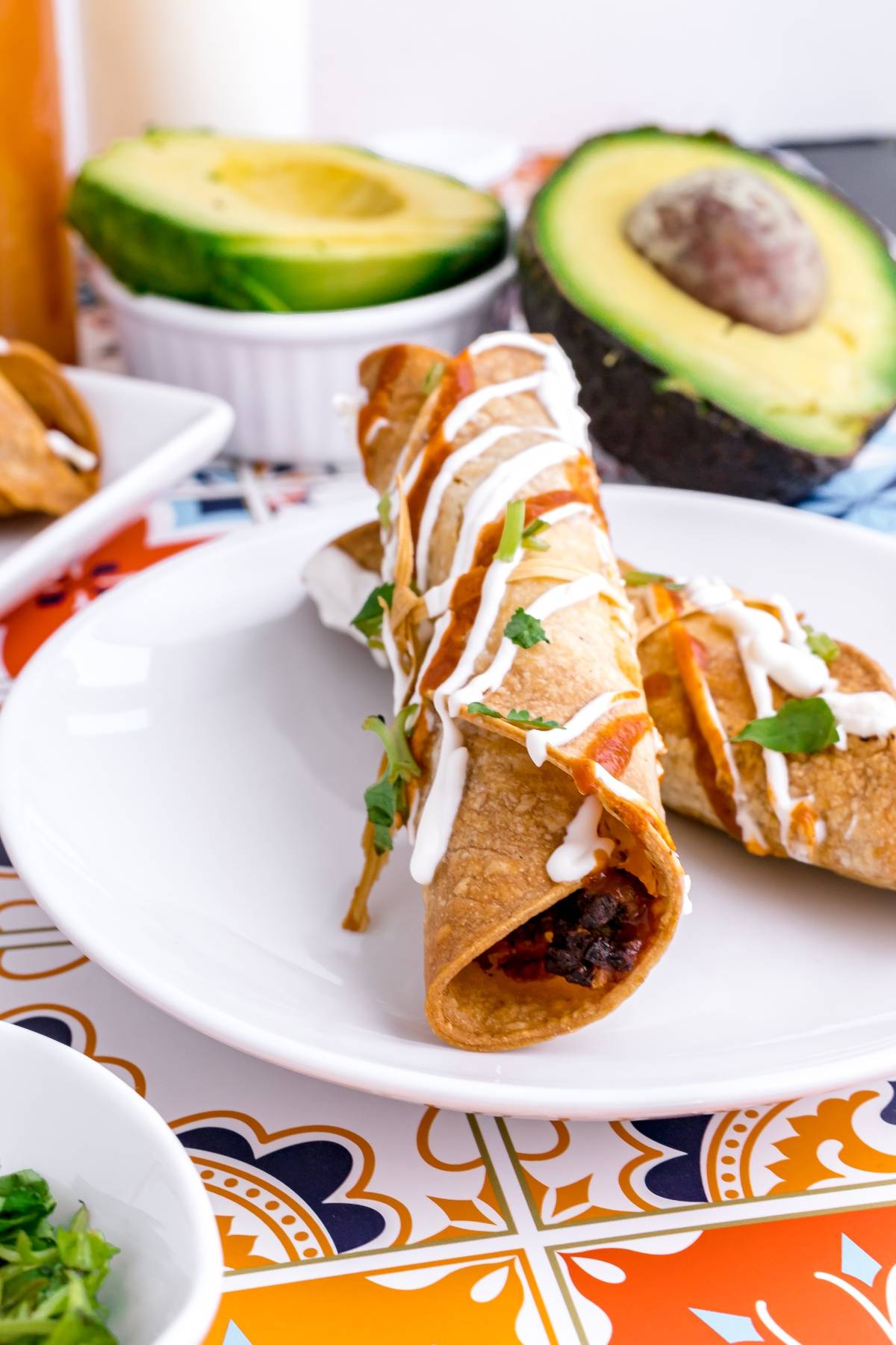 Two crispy air fryer taquitos drizzled with white sauce on a plate, with sliced avocado in the background—a delicious option for your next gluten free Mexican dinner.