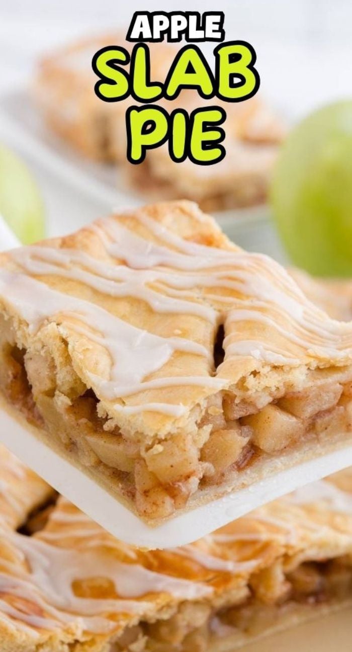 A slice of apple slab pie with icing drizzle is being lifted, while fresh green apples sit in the background.