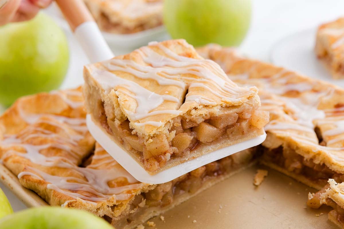 A spatula lifts a square slice of glazed apple slab pie bars from a baking pan, with apples in the background.