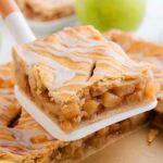 A spatula lifts a square slice of glazed apple slab pie from a baking pan, with green apples in the background.