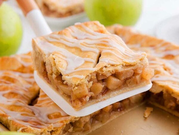 A spatula lifts a square slice of glazed apple slab pie from a baking pan, with green apples in the background.