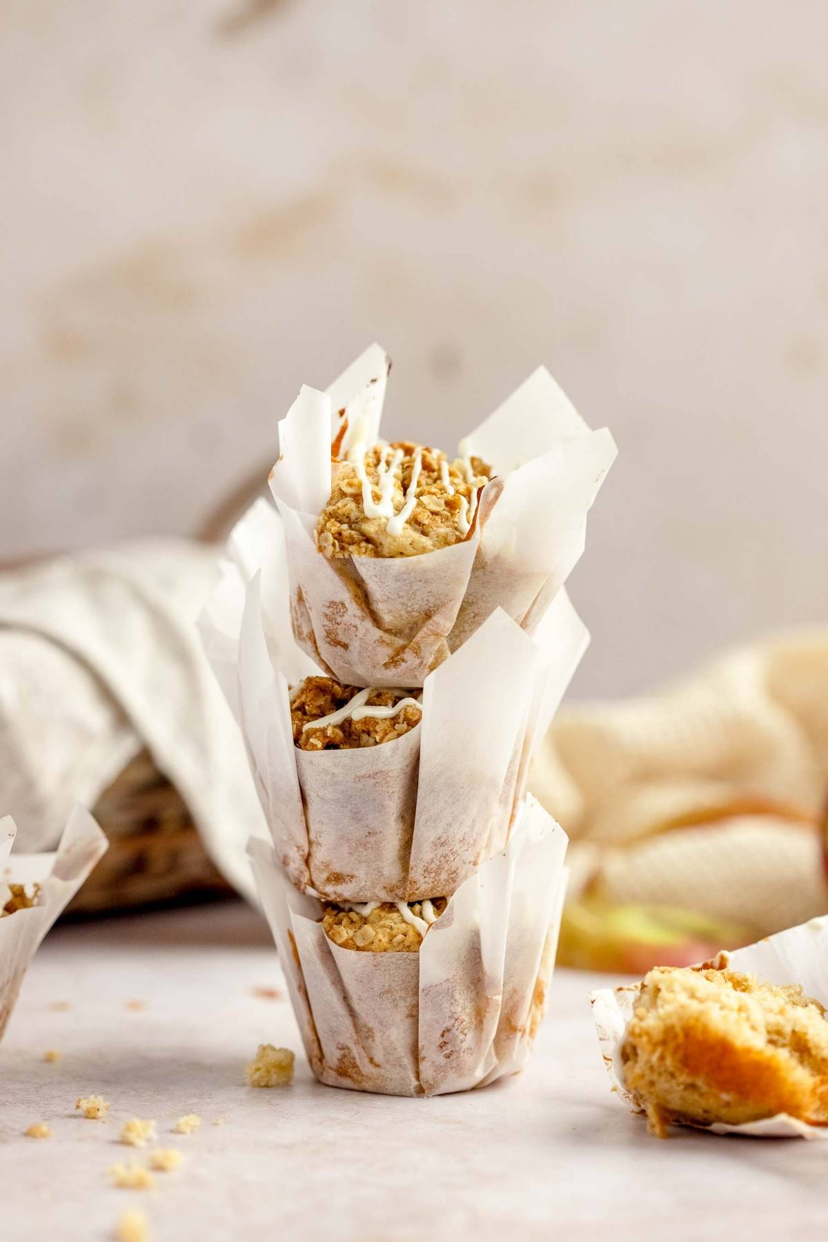 Three Apple Cinnamon Crumb Muffins stacked in white parchment wrappers on a light surface, with crumbs scattered around.