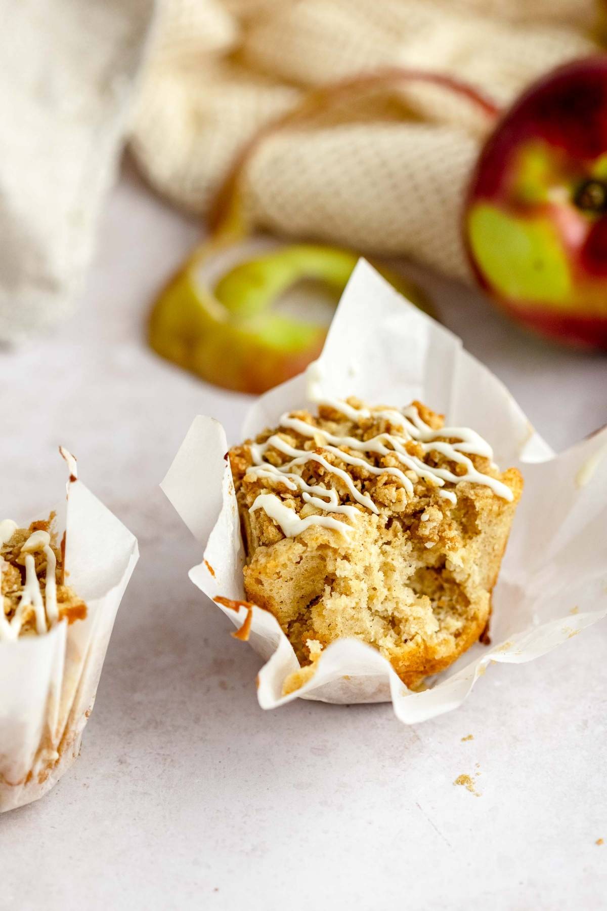 A partially eaten autumn apple muffin with white icing sits in a paper wrapper, with an apple and peel in the background.