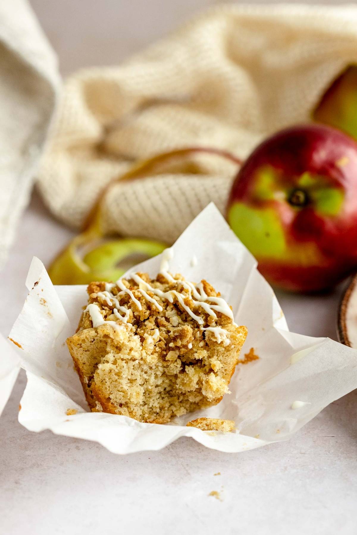 A partially eaten Apple Cinnamon Crumb Muffin with icing sits among apples and a beige cloth in the background.