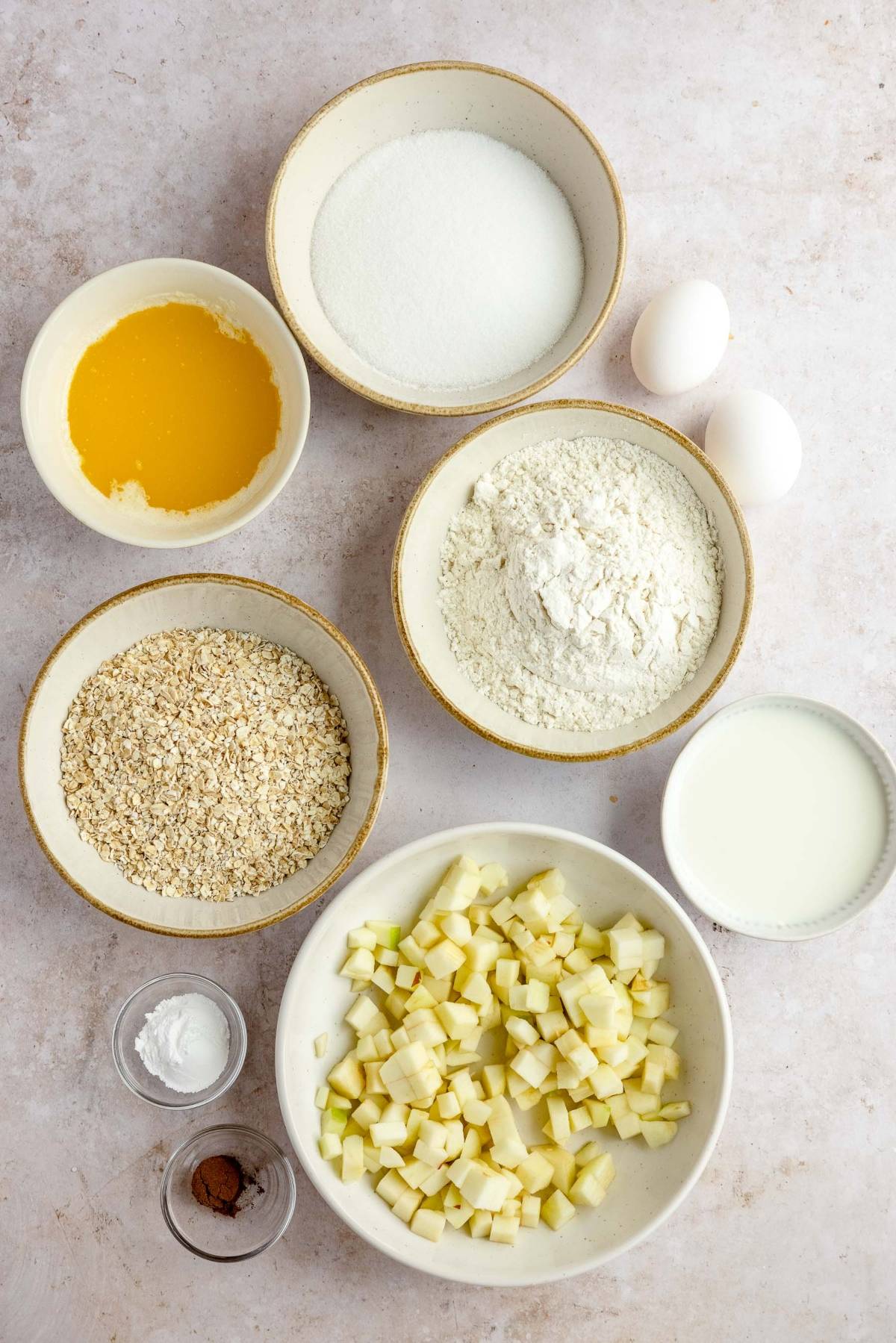 Bowls of diced apples, oats, flour, sugar, melted butter, milk, baking powder, cinnamon, and two eggs on a countertop—perfect for whipping up an autumn apple muffins or a delicious apple crumble muffins recipe.