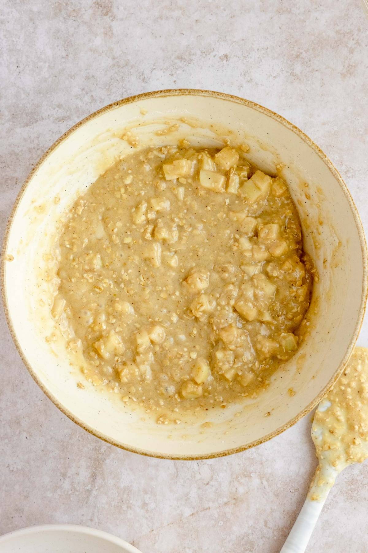 Bowl of lumpy, beige batter with chunks for apple oatmeal muffins with streusel topping, and a spoon resting nearby on a light surface.