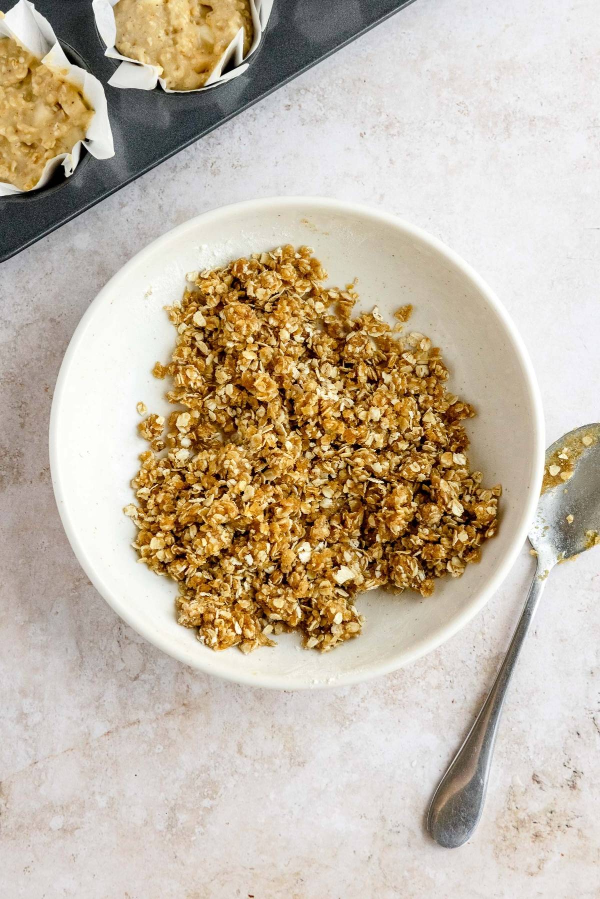 A white bowl with oat crumble mixture beside a spoon, near a muffin tin filled with batter in paper liners, ready to bake delicious apple oatmeal muffins with streusel topping.