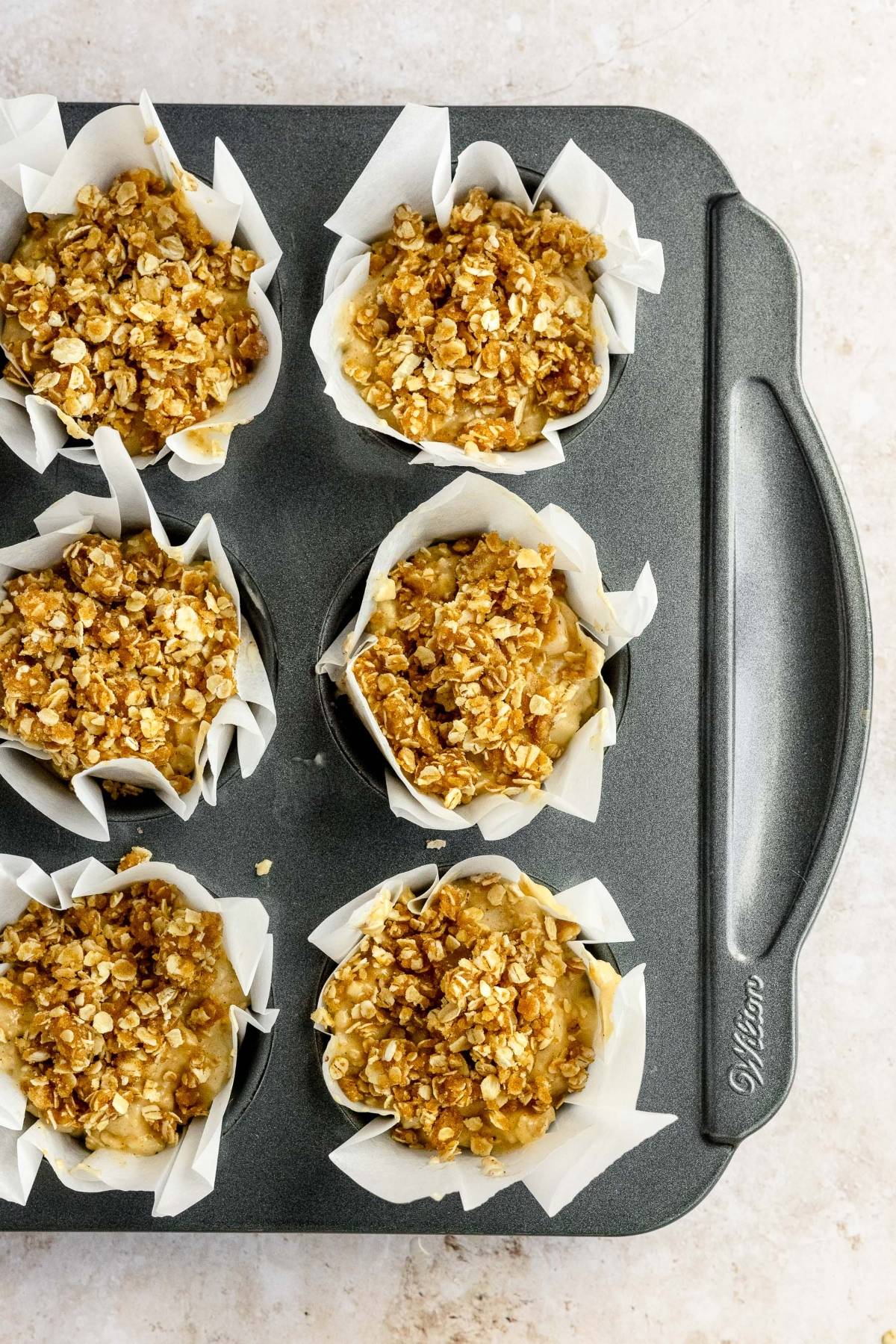 Six muffin cups in a pan, filled with oat crumble topping and lined with parchment paper, ready to bake this delicious apple crumble muffins recipe.