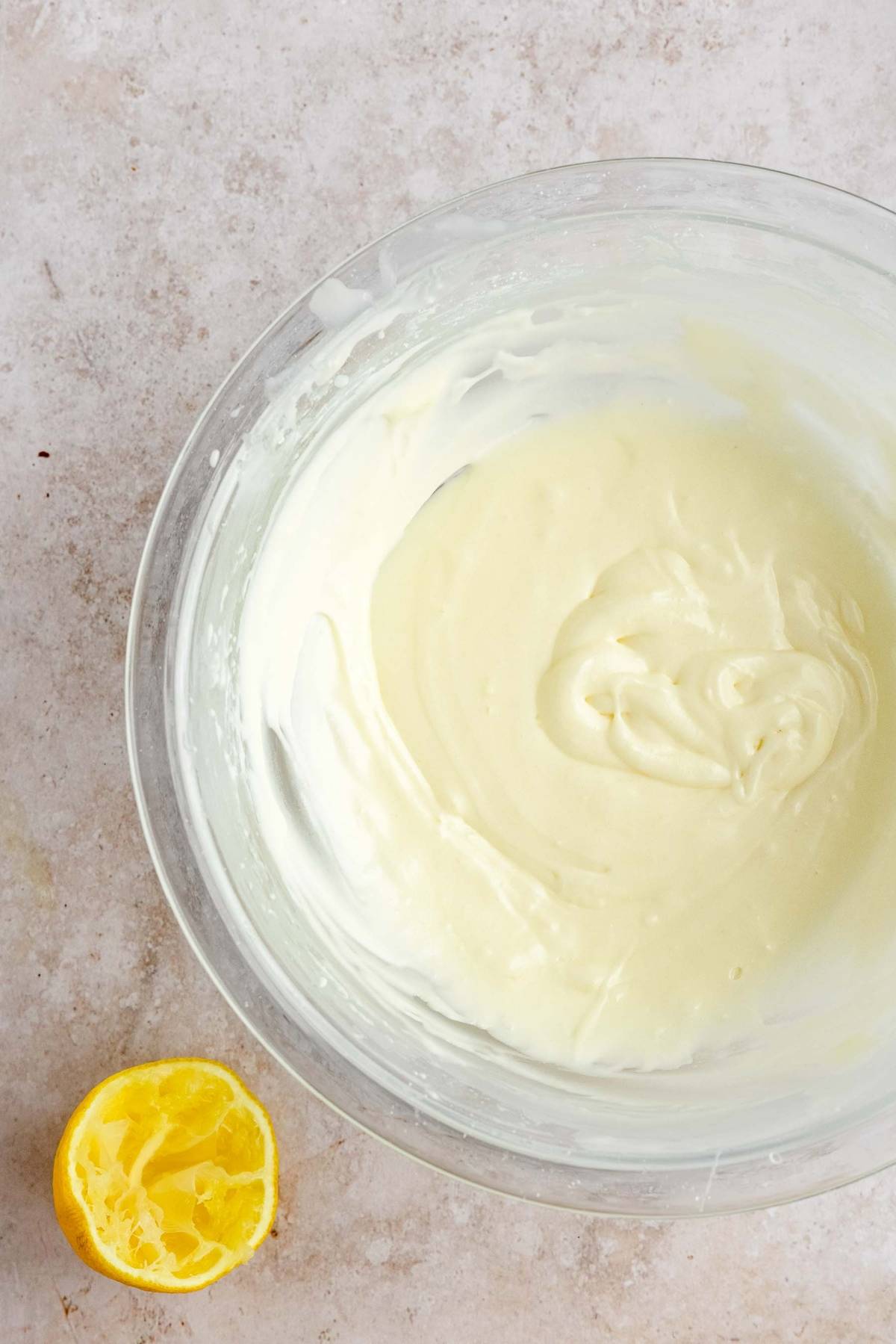 A glass bowl of creamy mixture beside a squeezed half lemon on a light countertop, perfect for whipping up an apple crumble muffins recipe.