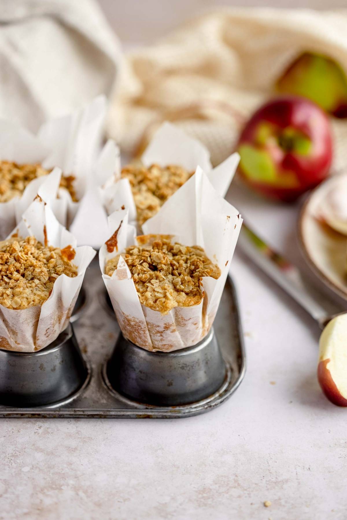 Apple crumble muffins in parchment paper in a muffin tin, with apples and a plate in the background—an inviting autumn apple muffins treat perfect for cozy days.