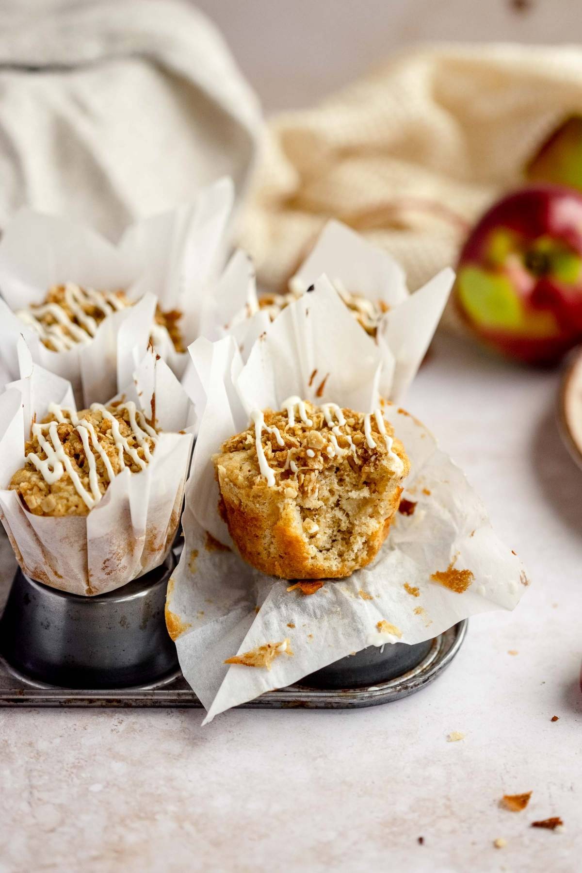 Apple Cinnamon Crumb Muffins with white icing sit in parchment wrappers, one partially unwrapped in a muffin tin—perfectly capturing the cozy flavors of autumn apple muffins.