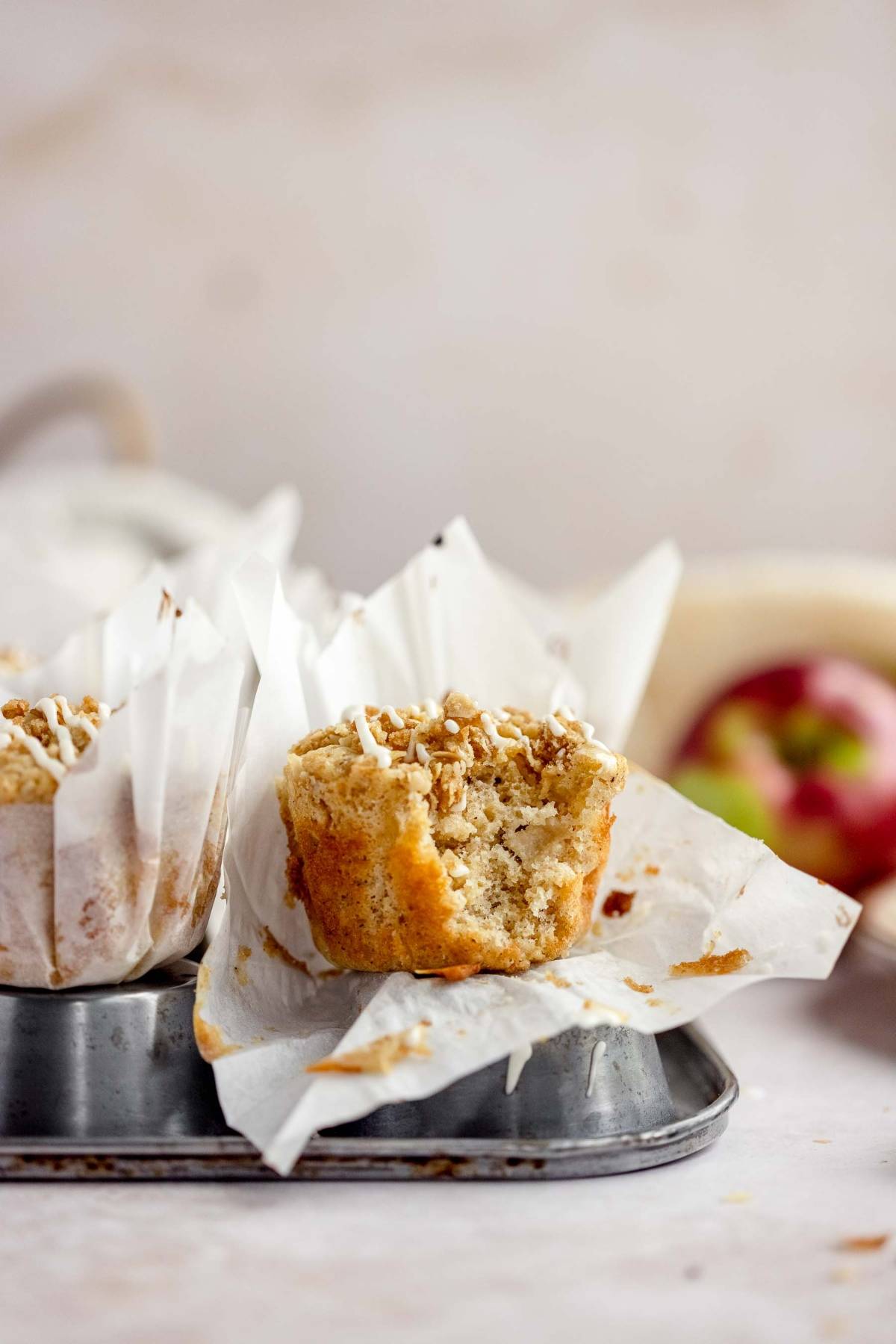 A muffin with a bite taken out, topped with crumbs, sits in a baking tray lined with white parchment paper—just like a classic Apple Cinnamon Crumb Muffin.