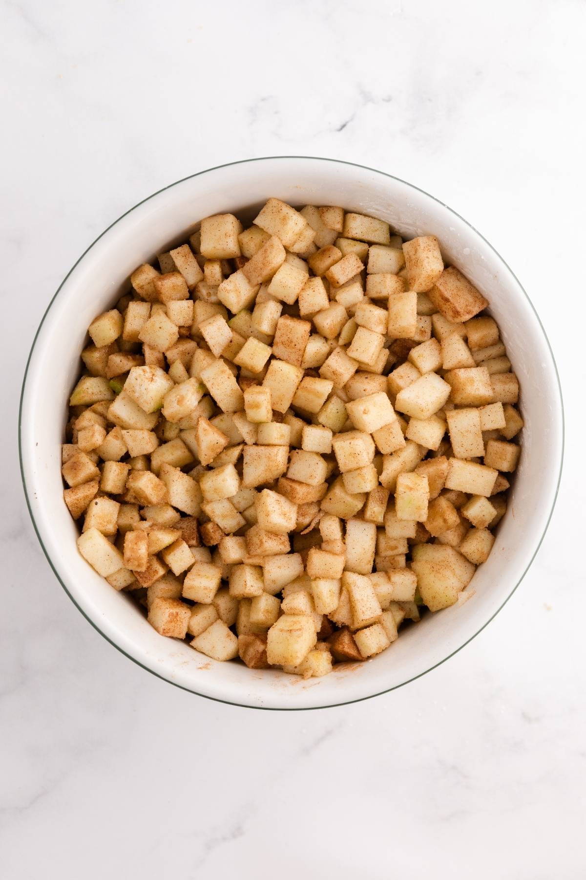 A bowl filled with small diced apples coated in cinnamon on a white marble surface.