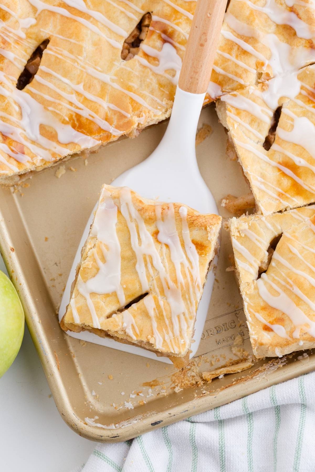 A glazed square of apple slab pie is lifted from a baking pan with a spatula.