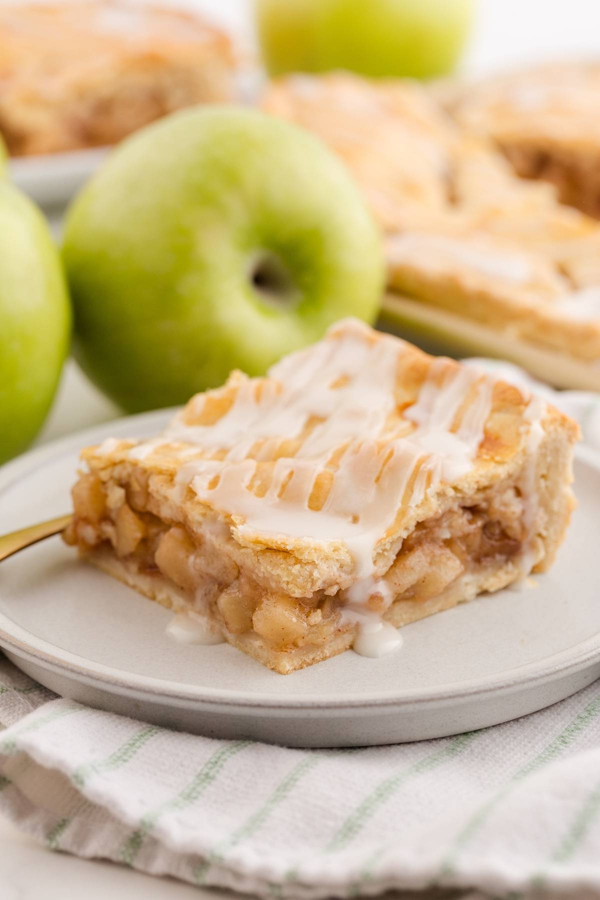 A slice of apple slab pie with icing on a plate, next to green apples and a striped cloth.