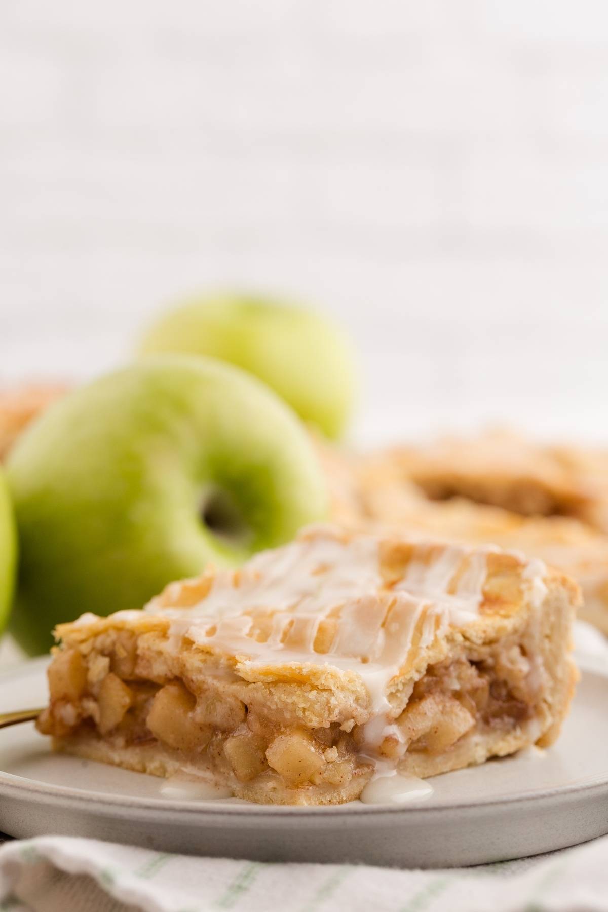 A slice of glazed apple sheet pan pie on a plate, with green apples in the background.