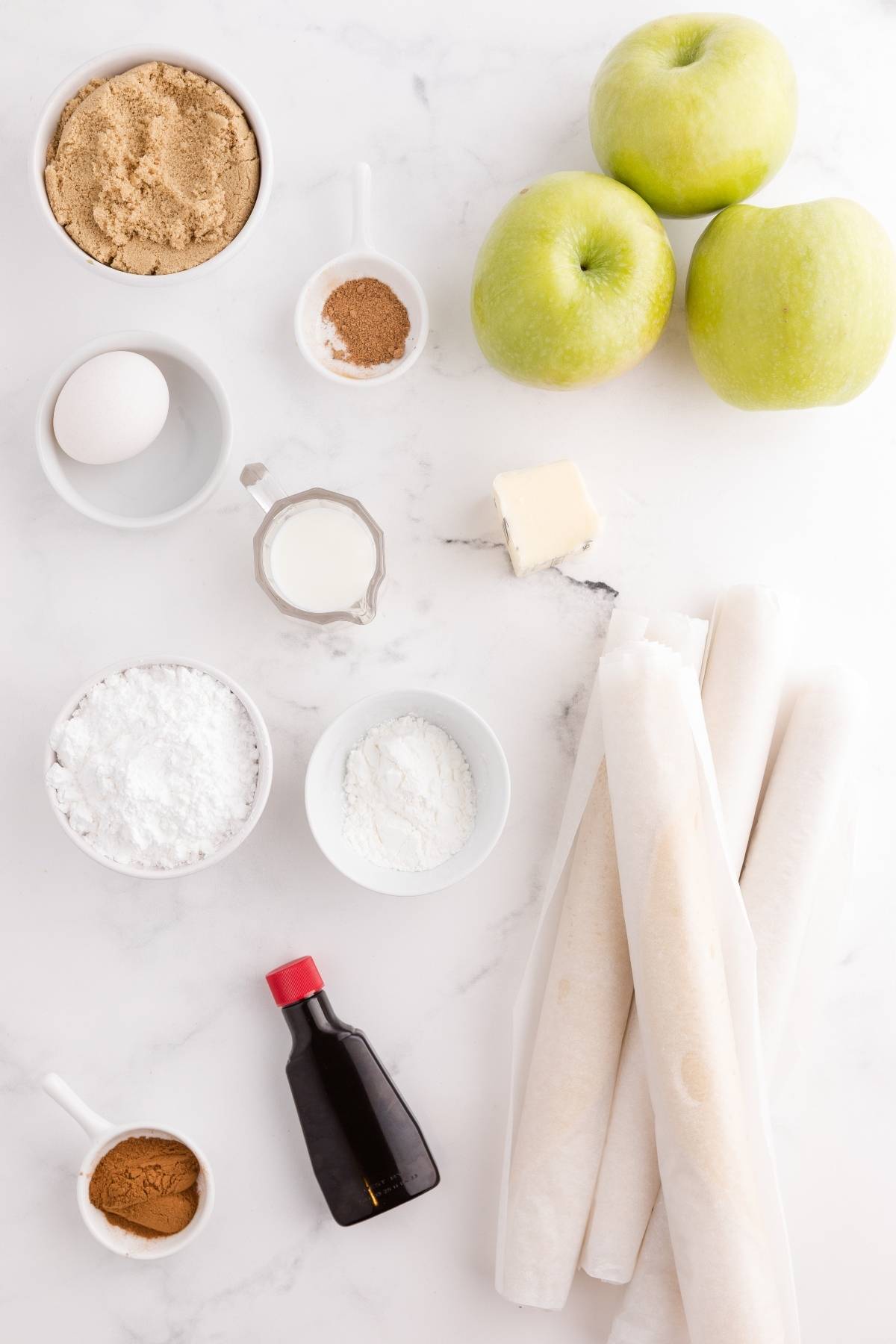Baking ingredients and green apples arranged on a white surface, including flour, sugar, cinnamon, and pie crusts.