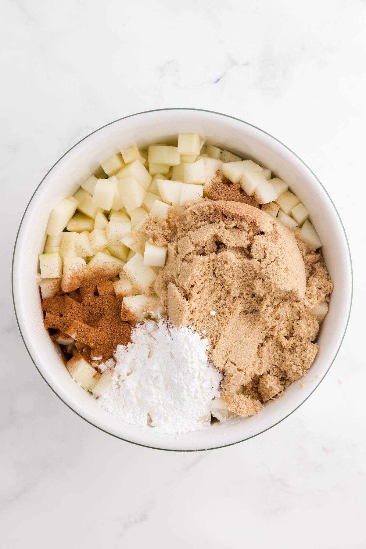 White bowl with chopped apples, brown sugar, cinnamon, and cornstarch on a white marble surface.