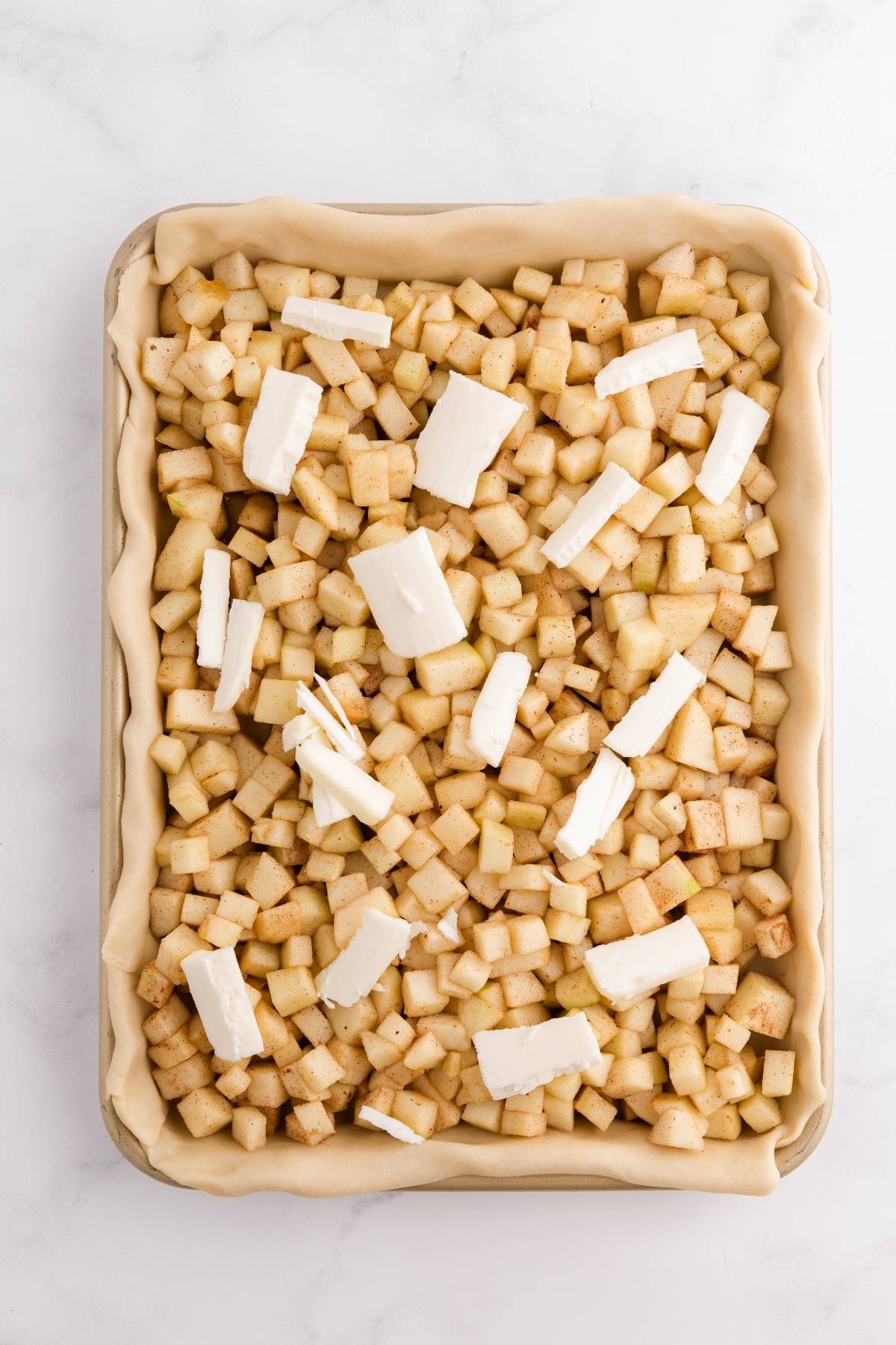 Unbaked pie with diced apples, cinnamon, and butter pieces on a crust in a rectangular baking pan.