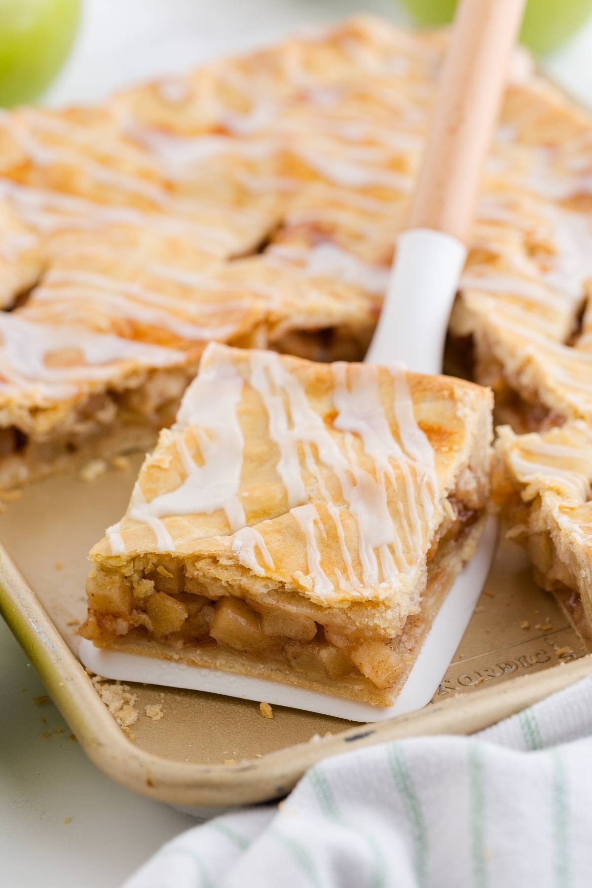 A spatula lifts a slice of glazed apple pie bar from a baking pan, showing the apple filling inside.
