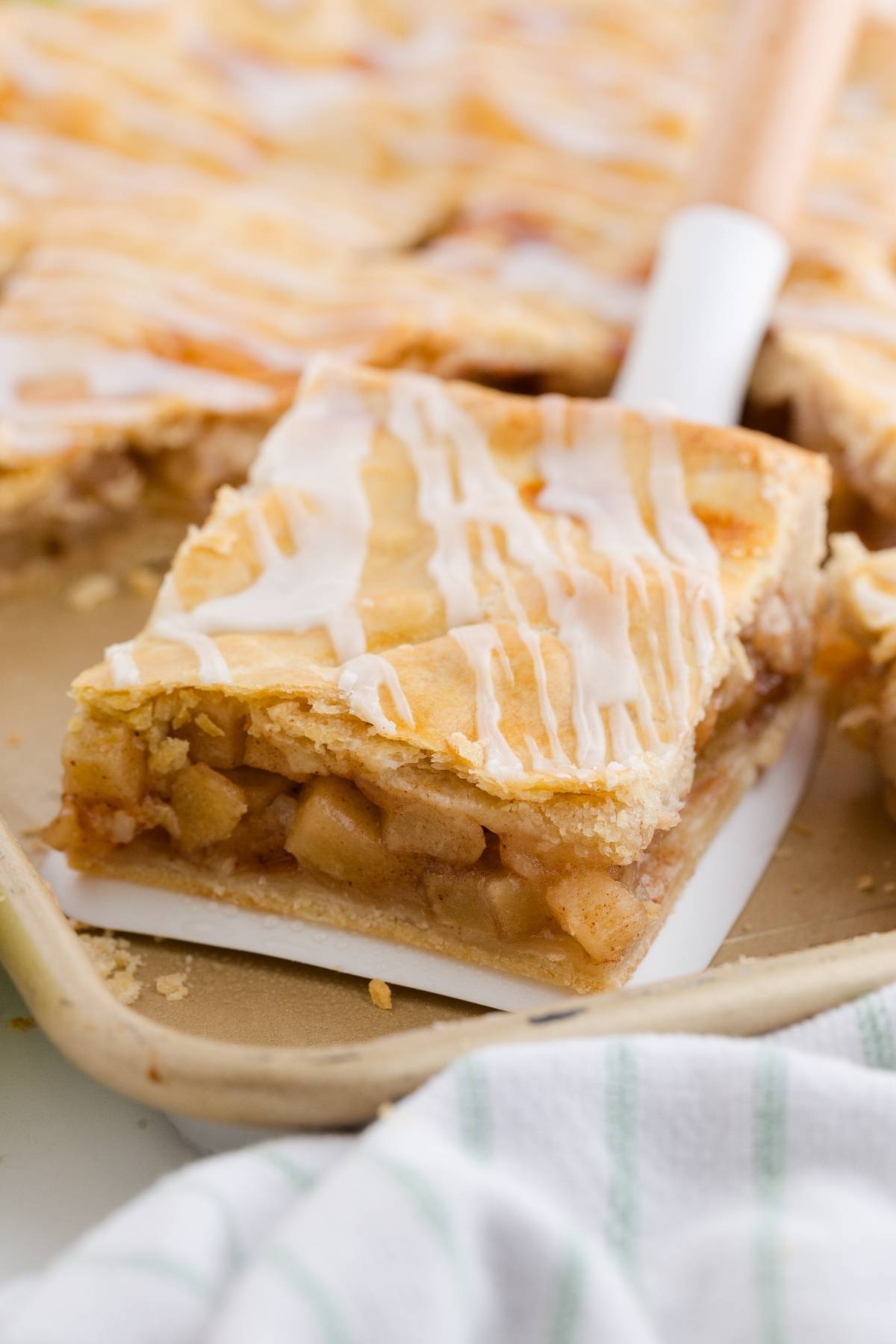 A square slice of glazed apple slab pie with a flaky crust on a spatula, showing apple filling.