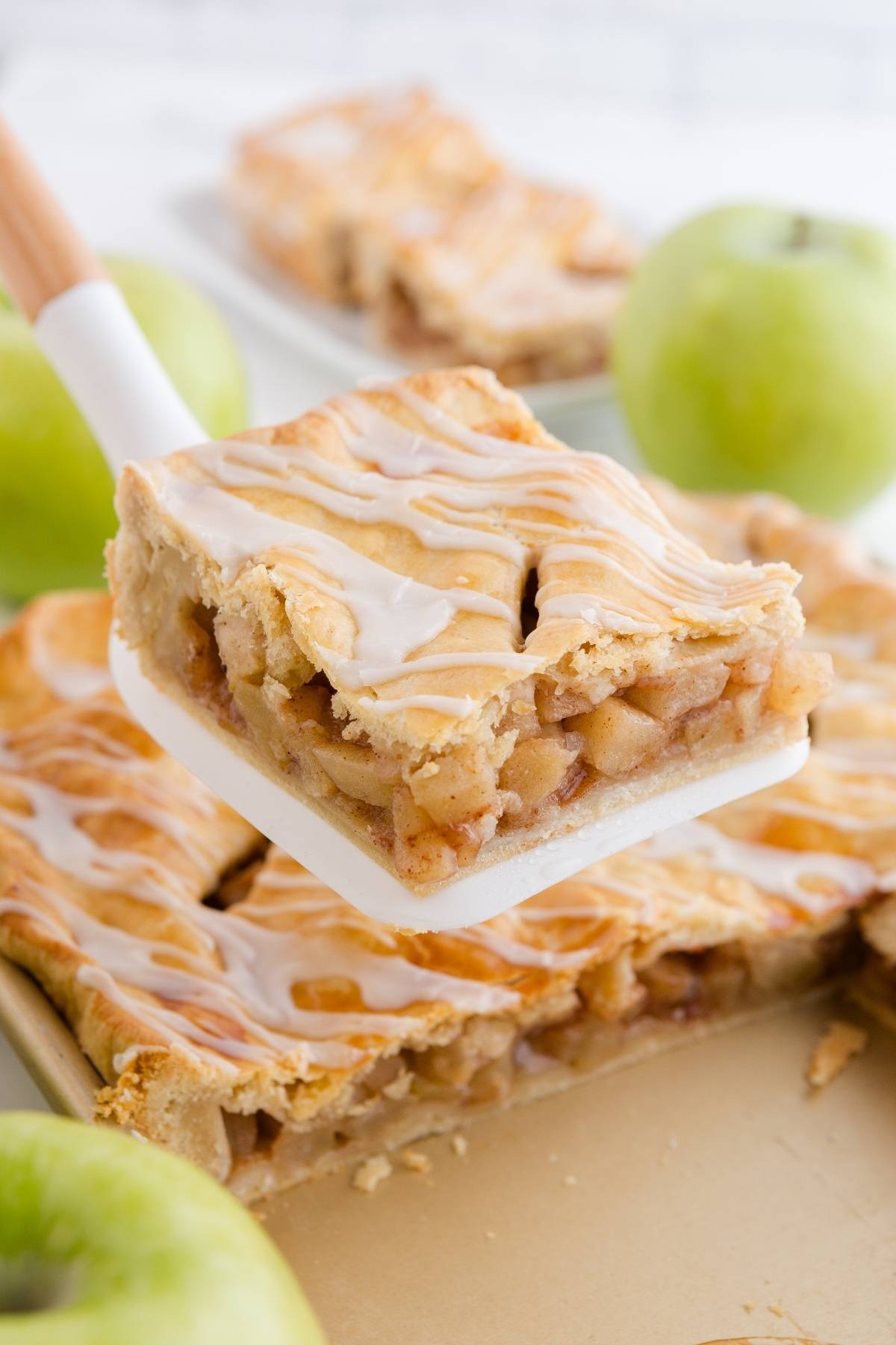 A spatula holds a slice of glazed apple pie, with fresh green apples in the background.