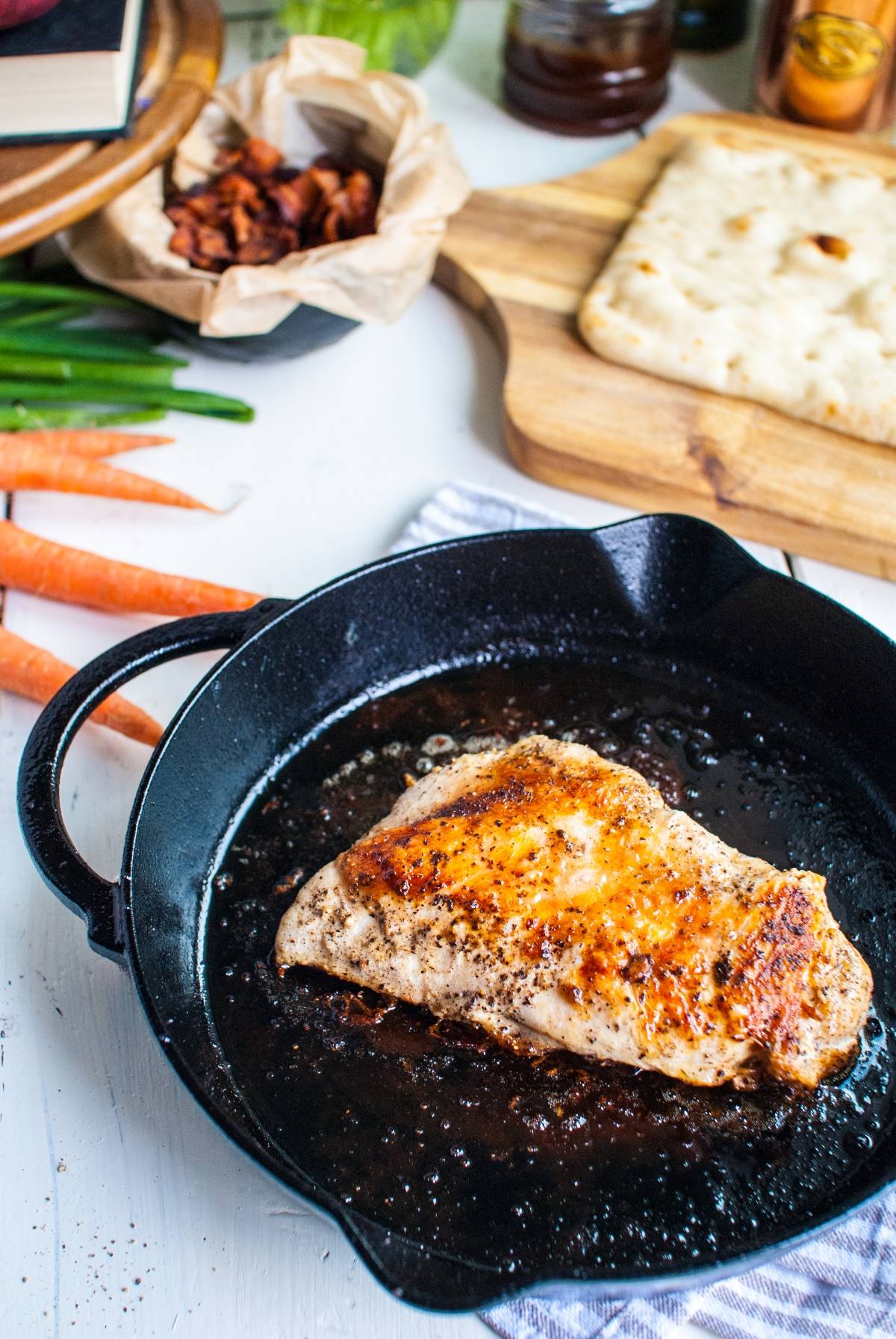 Seasoned chicken breast cooking in a black skillet, perfect for easy BBQ chicken pizza, with vegetables and bread on a wooden board nearby.