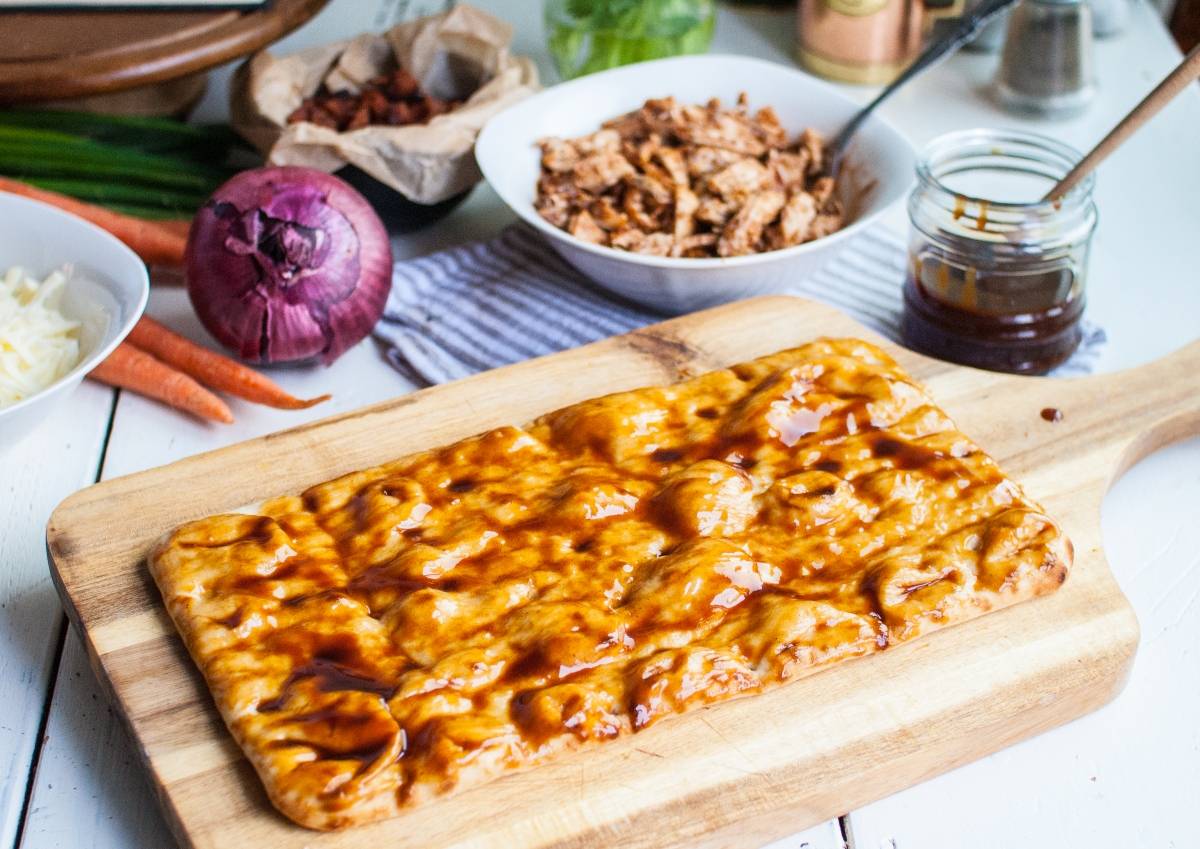 Rectangular pizza crust on a wooden board, topped with barbecue sauce—an easy BBQ chicken pizza made in under 30 minutes; fresh ingredients surround it.