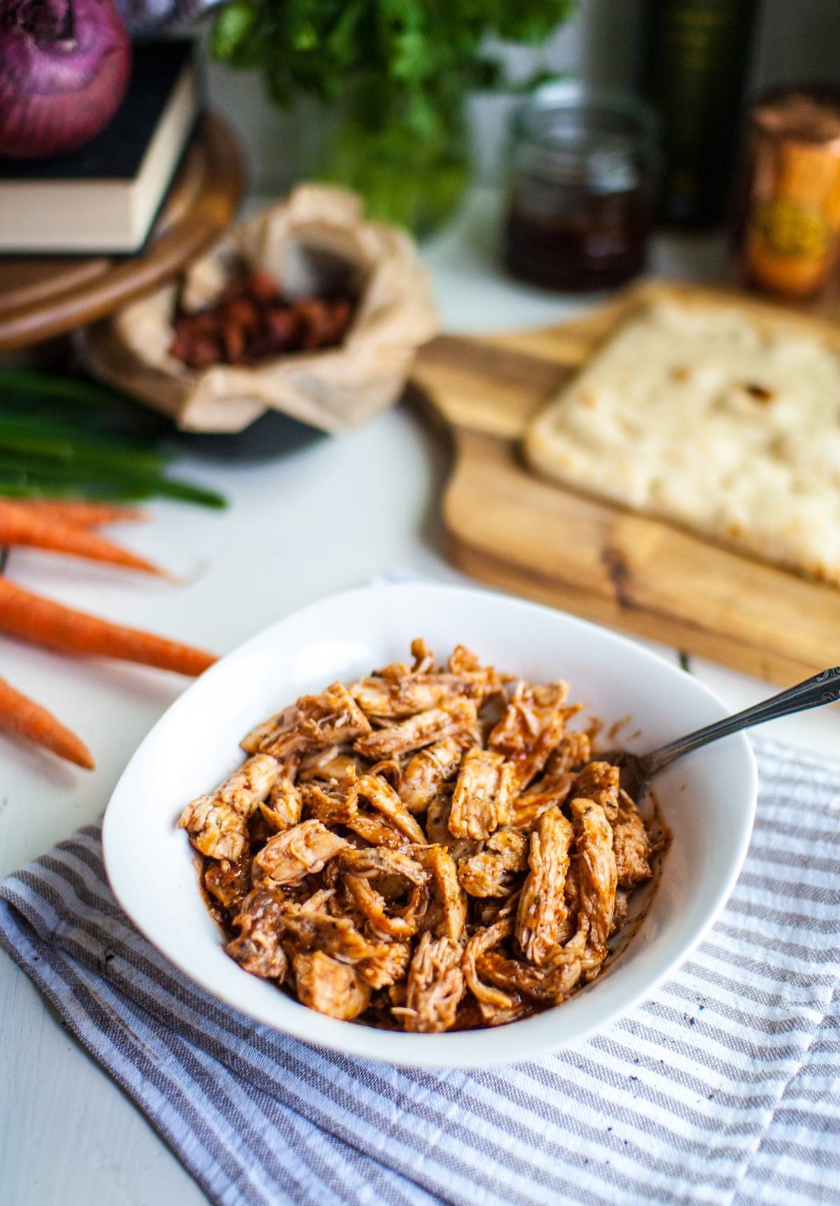 Shredded barbecue chicken in a white bowl with a fork, surrounded by veggies and flatbread on a table—perfect for making barbecue chicken flatbread like Panera at home.
