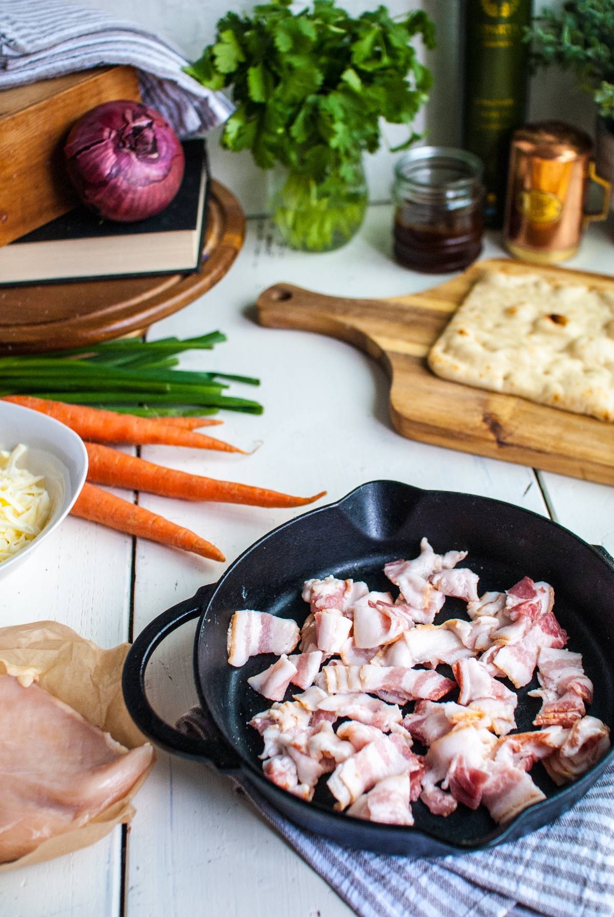 Chopped bacon in a cast iron skillet with carrots, flatbread, cheese, and herbs on a kitchen counter—perfect ingredients for an easy BBQ chicken pizza made in under 30 minutes.