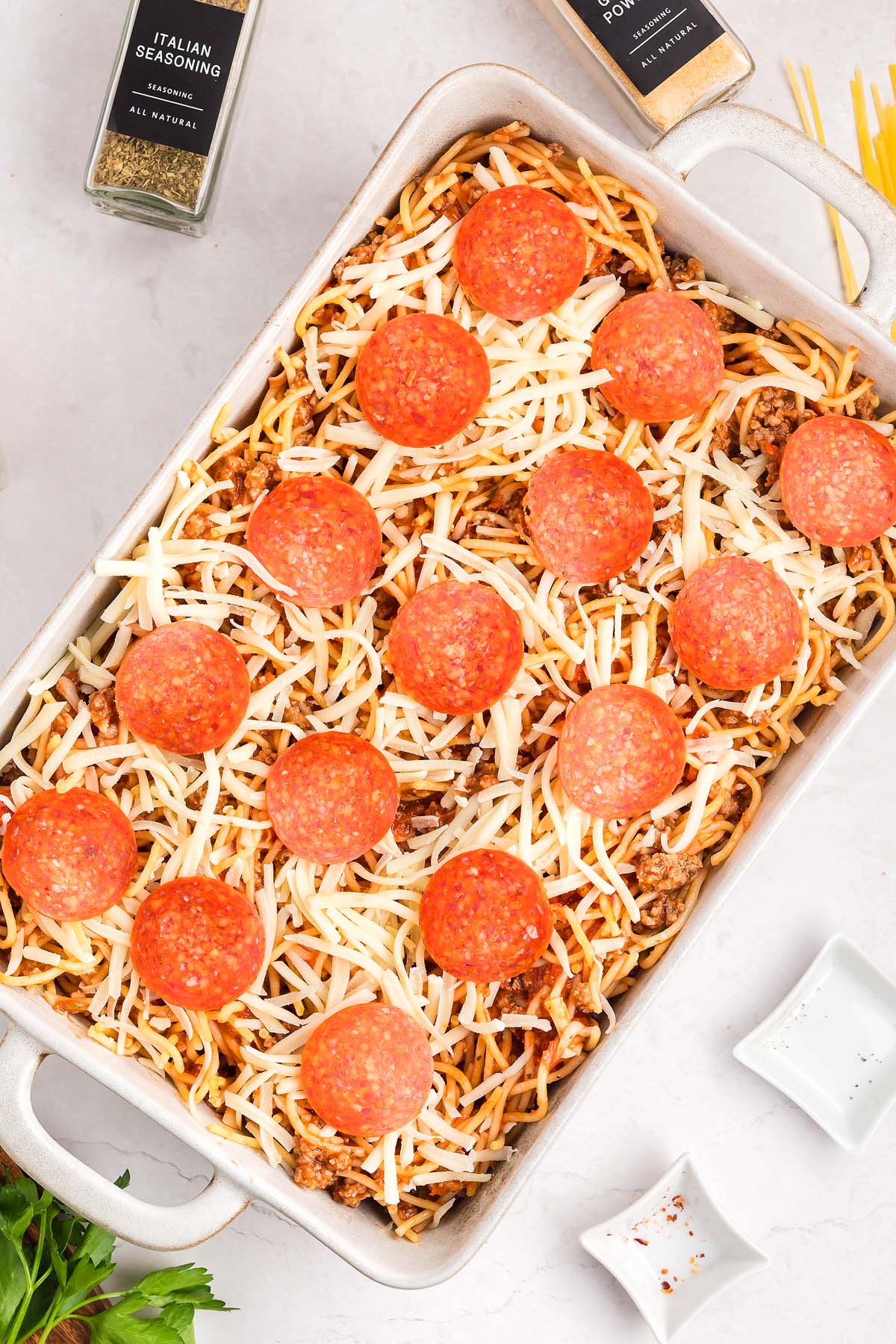 A casserole dish of baked spaghetti pizza, topped with shredded cheese and pepperoni slices, ready to be cooked.