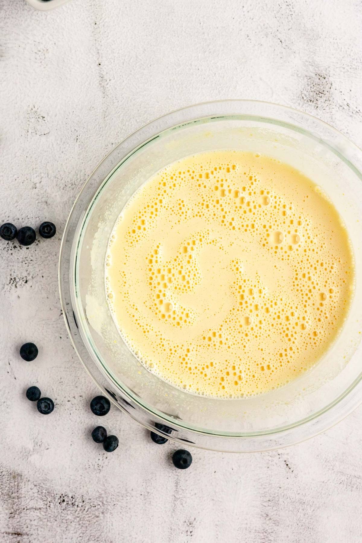 A glass bowl of pale yellow batter sits on a light-colored surface, with blueberries scattered nearby—perfect for making homemade Blueberry Cheesecake Ice Cream.