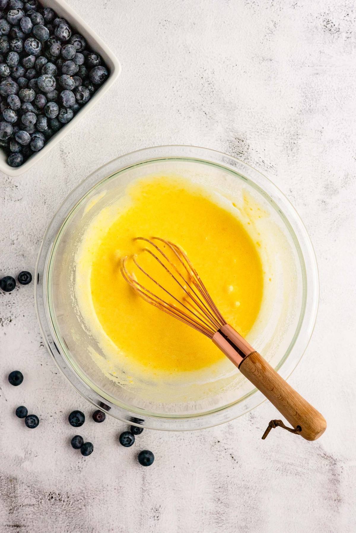 A glass bowl of yellow batter with a whisk sits beside a bowl of blueberries and scattered berries—perfect for making Blueberry Cheesecake Ice Cream, made with fresh blueberries and cream cheese.