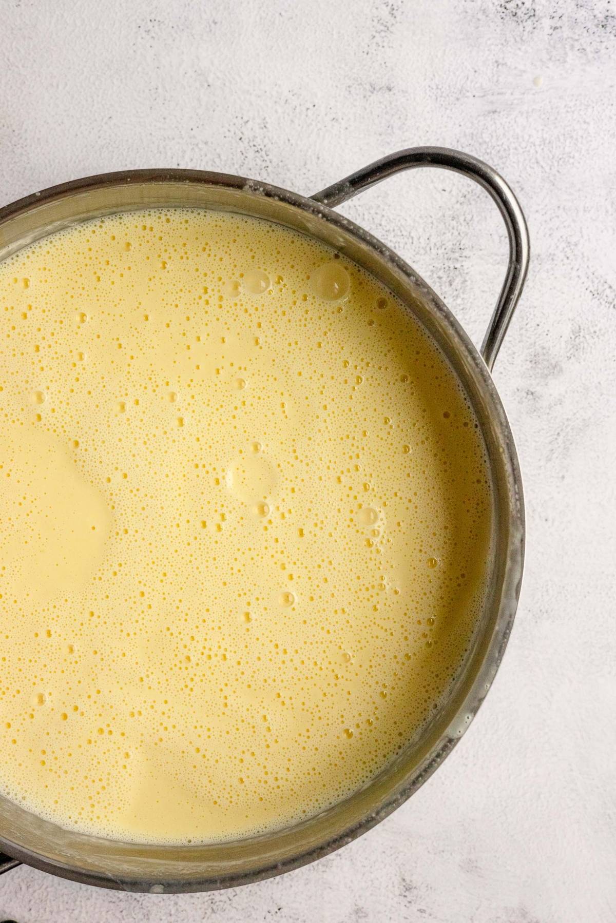 A metal pot filled with a yellow, frothy liquid on a light-colored textured surface, perfect for starting a homemade cheesecake ice cream.