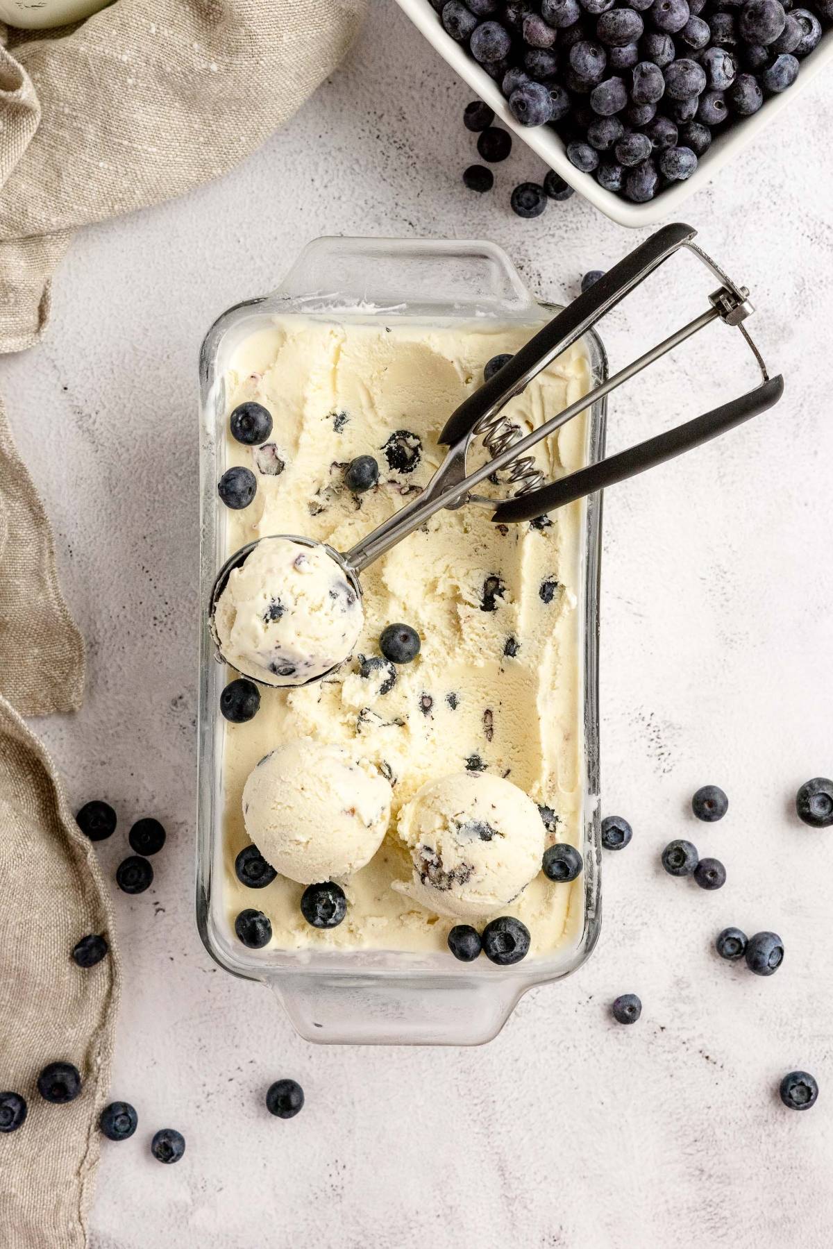 Vanilla ice cream with blueberries in a glass dish, scooped with an ice cream scooper, surrounded by fresh blueberries—a perfect blueberry cheesecake ice cream treat made with fresh blueberries and cream cheese.