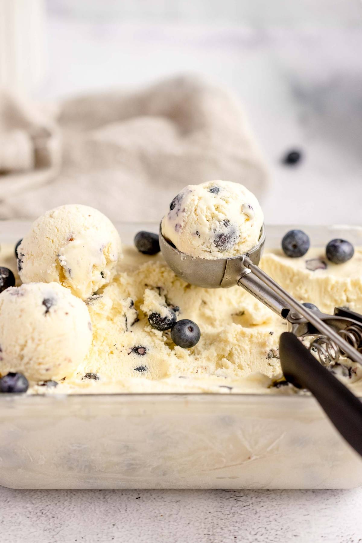 Scoops of blueberry cheesecake ice cream in a container, made with fresh blueberries and cream cheese, served alongside an ice cream scooper.