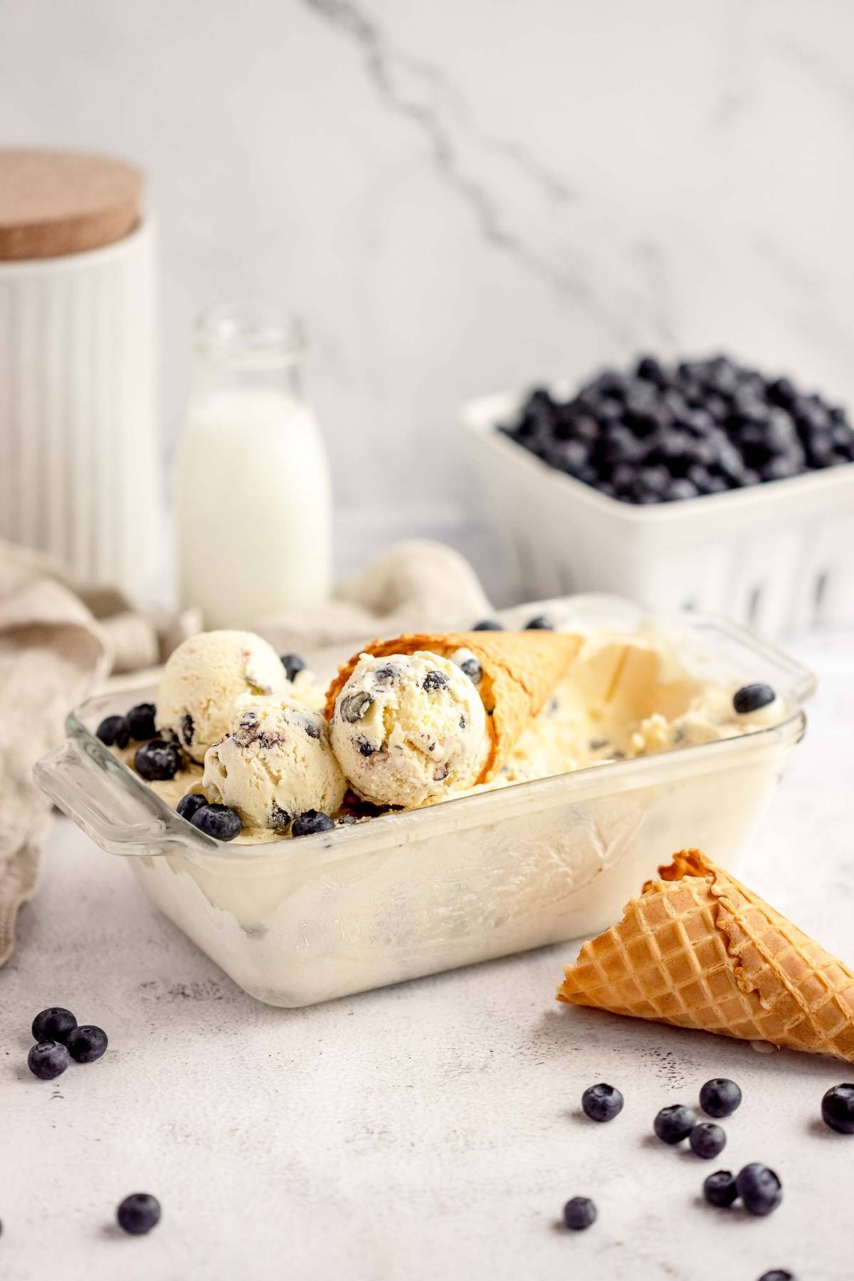 Vanilla ice cream with blueberries in a glass dish, served alongside scoops and waffle cones on a white surface—a tempting treat inspired by Blueberry Cheesecake Ice Cream, made with fresh blueberries and cream cheese.