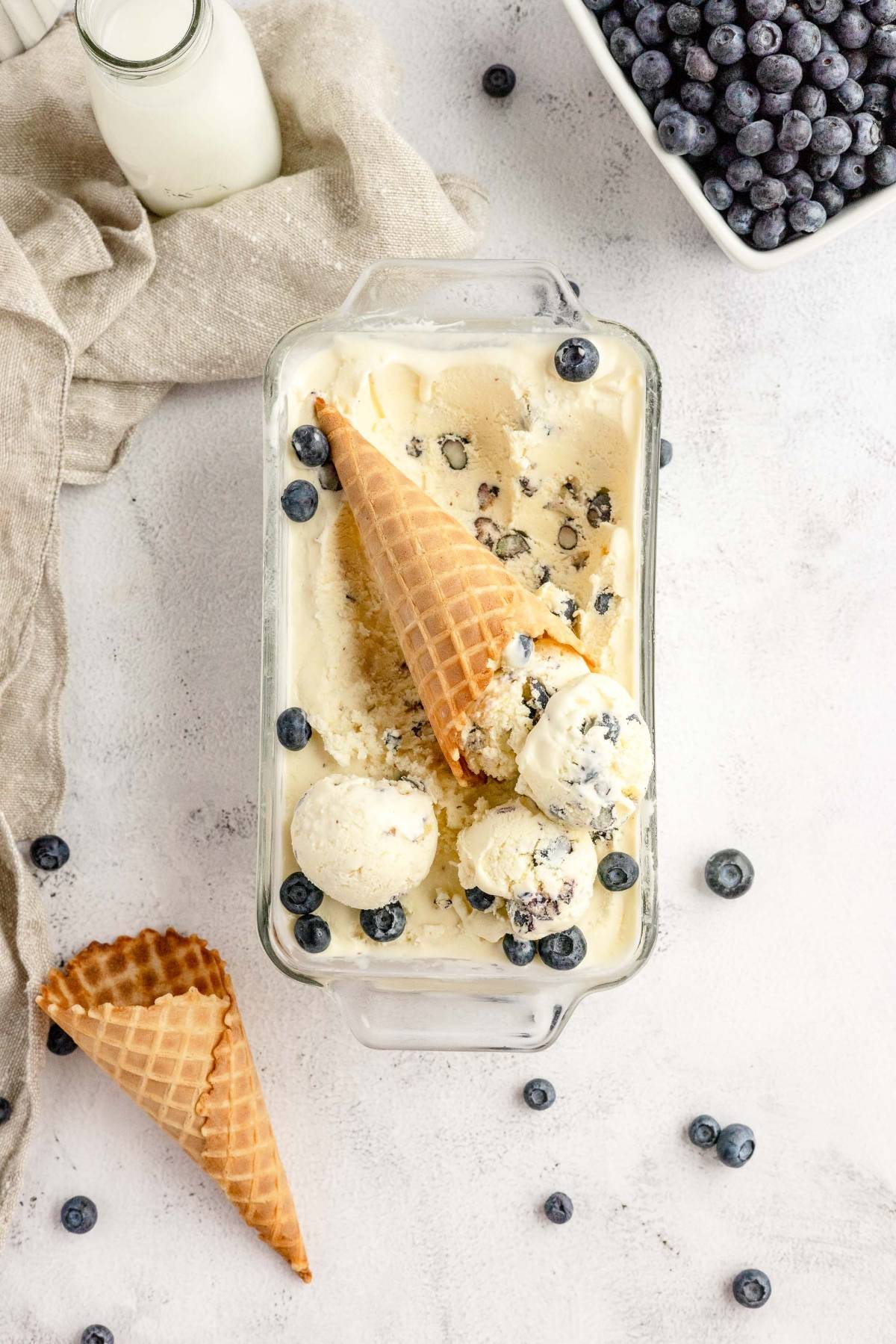 Glass dish of blueberry ice cream with waffle cones, milk, and fresh blueberries on a light surface; a homemade cheesecake ice cream made with fresh blueberries and cream cheese.