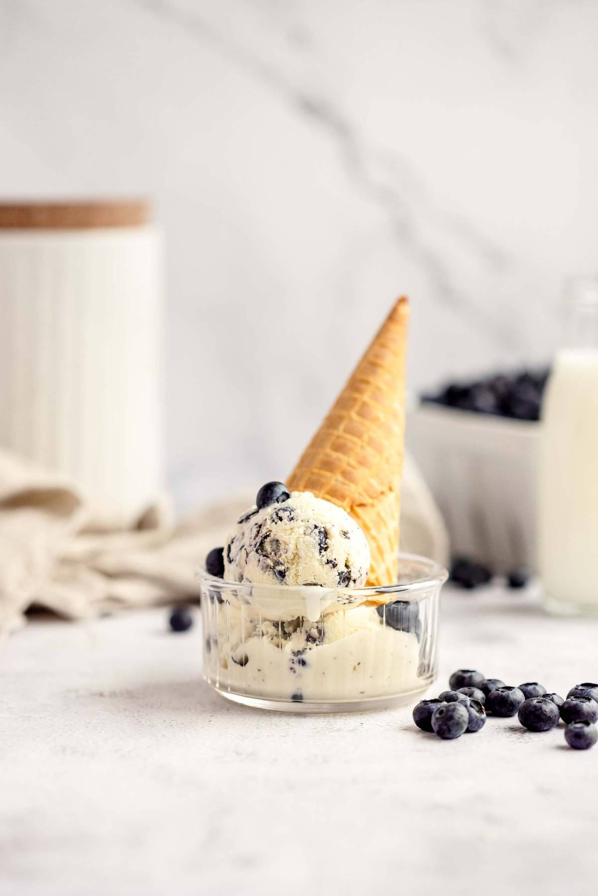 Ice cream cone with blueberries in a glass bowl, made with fresh blueberries and cream cheese, with milk and more blueberries in the background.