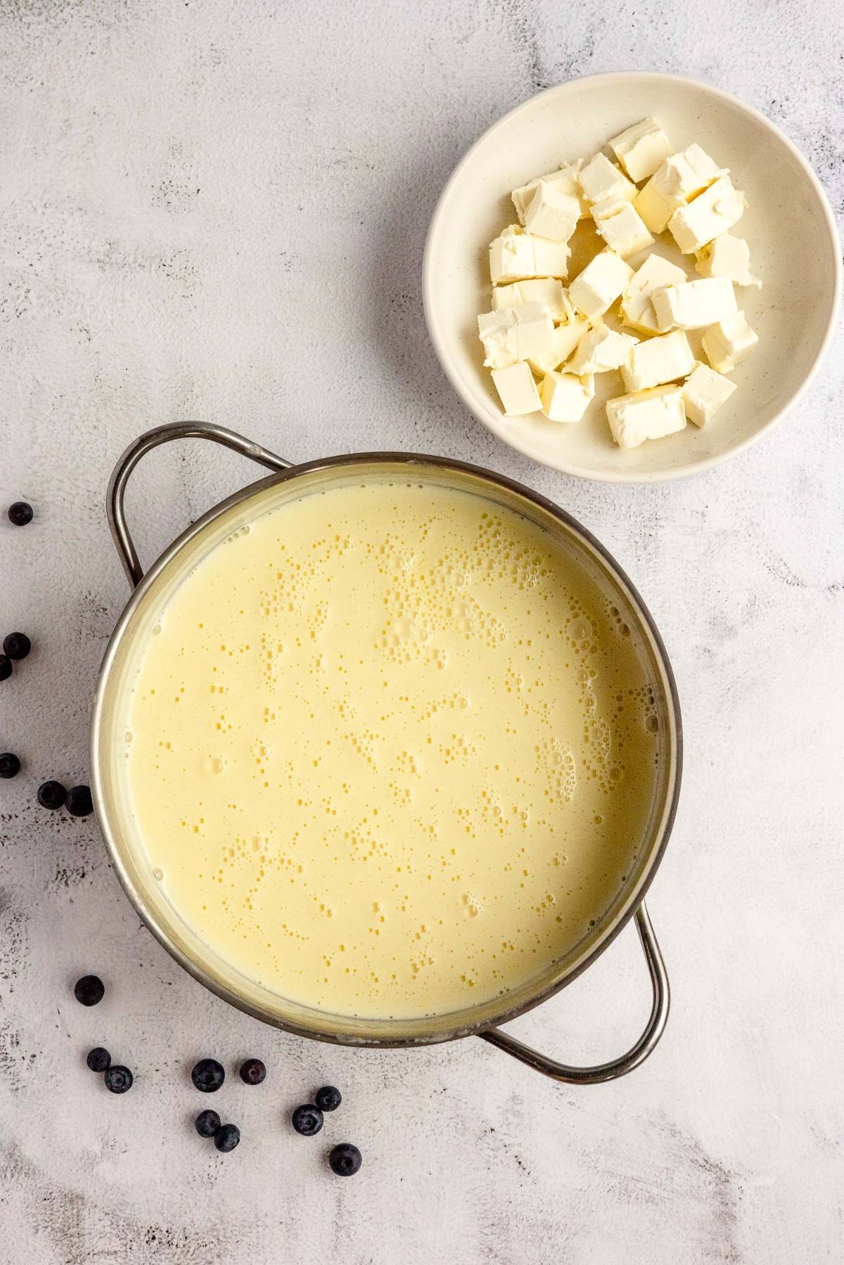 Large pot of creamy yellow mixture next to a bowl of cubed butter and scattered blueberries—perfect for a blueberry ice cream recipe made with fresh blueberries and cream cheese.