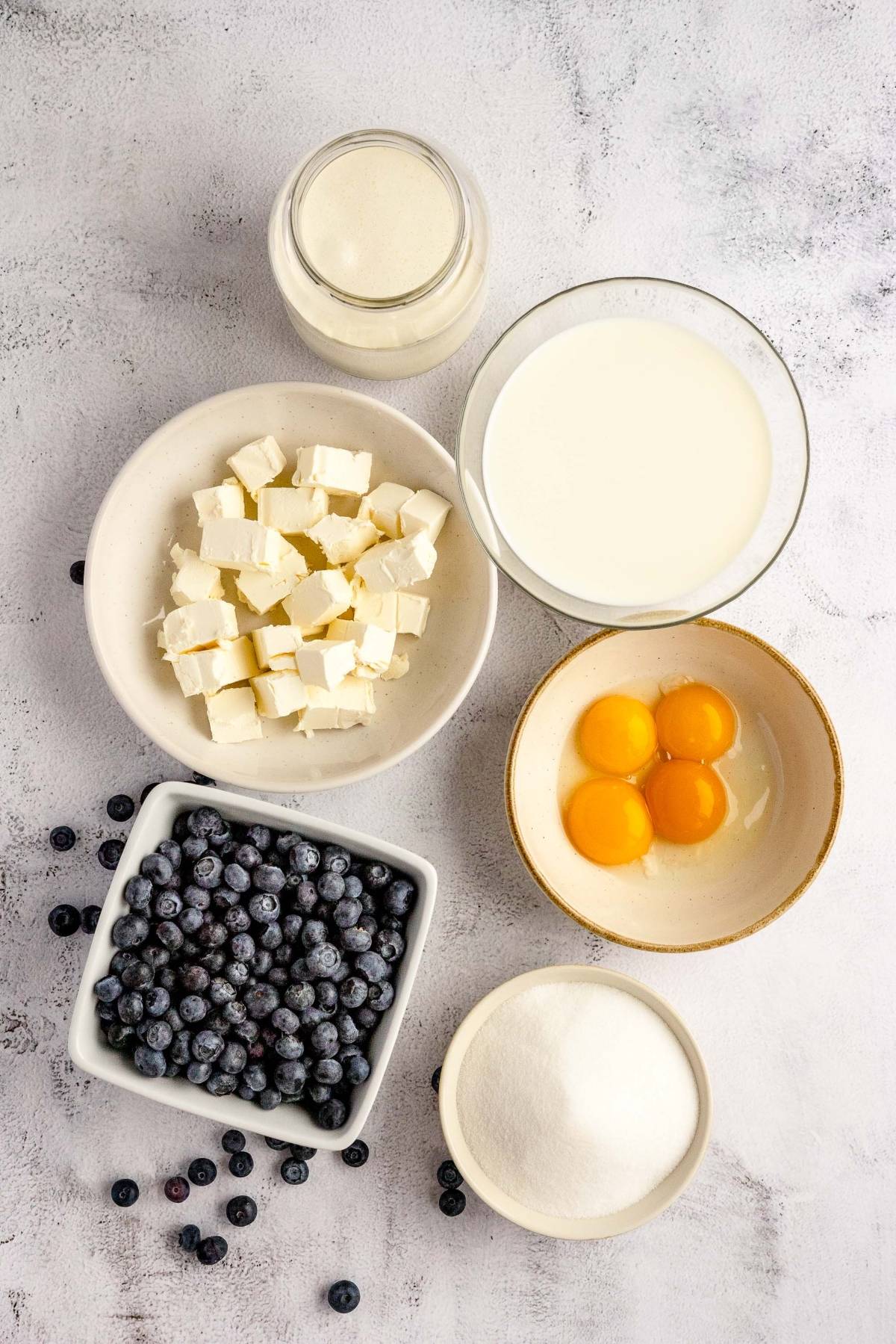 Bowls of blueberries, cubed cream cheese, eggs, sugar, and cream hint at homemade cheesecake ice cream made with fresh blueberries and cream cheese, all displayed on a light surface.