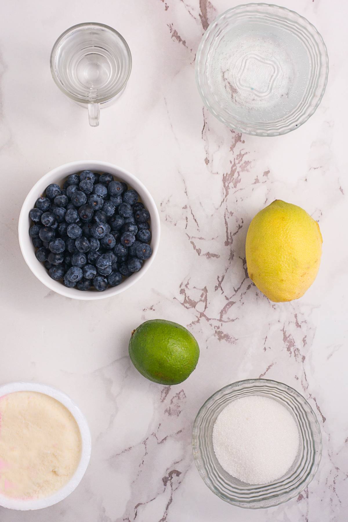 Ingredients on a marble surface: blueberries, lemon, lime, sugar, water, gelatin, and a small glass of liquid—perfect for crafting a sparkling blueberry float.